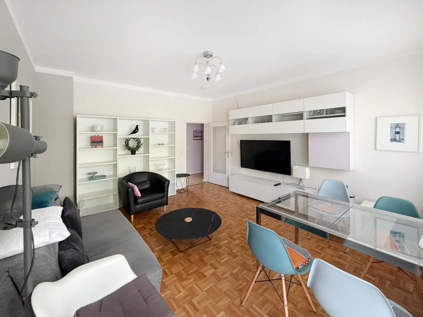 A bright living room with a gray sofa, black chair, glass table, and parquet floor. A white bookshelf and TV stand are against the wall.