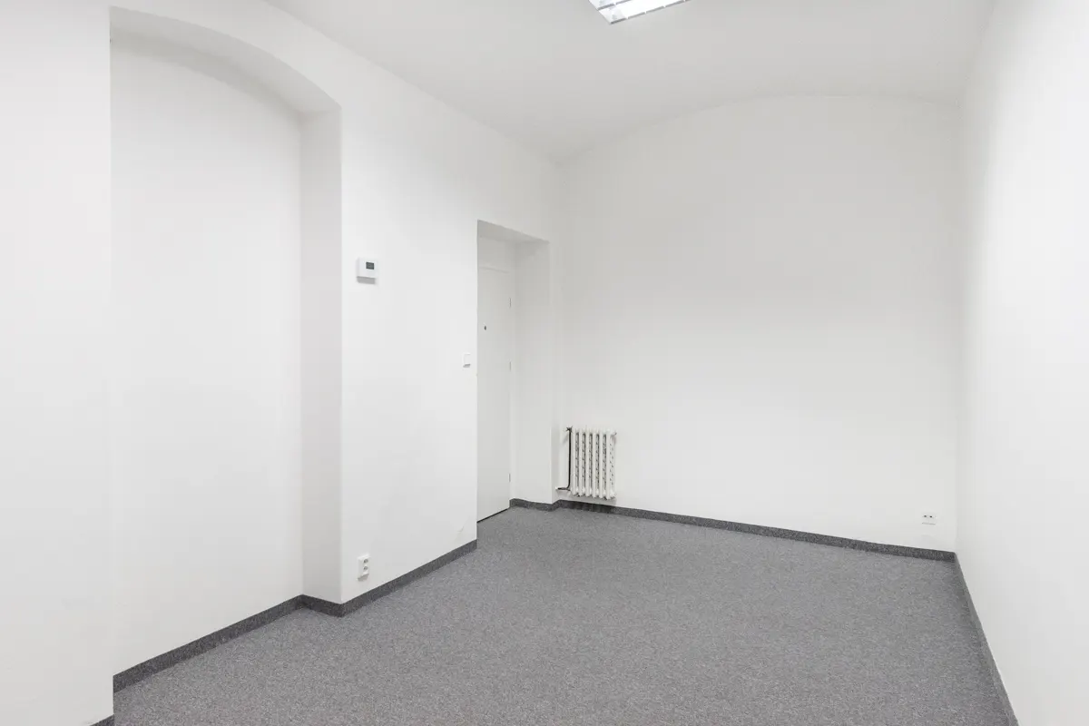 Empty room with white walls, gray carpet, and a radiator. A doorway is visible in the background.