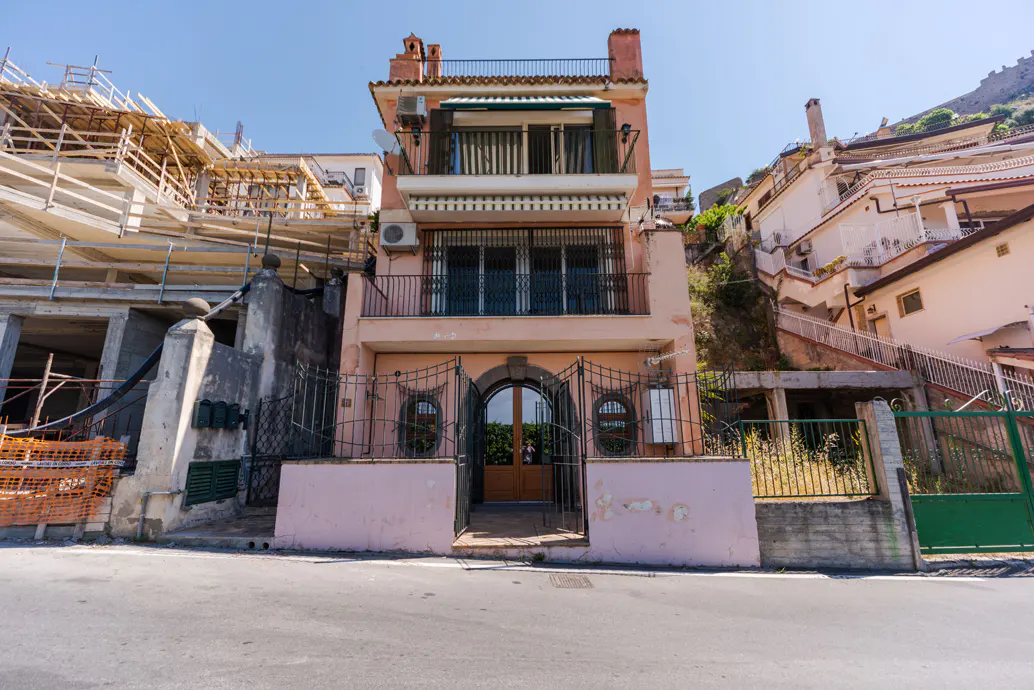 A three-story peach-colored house with balconies and a gated entrance, situated on a street next to a construction site.
