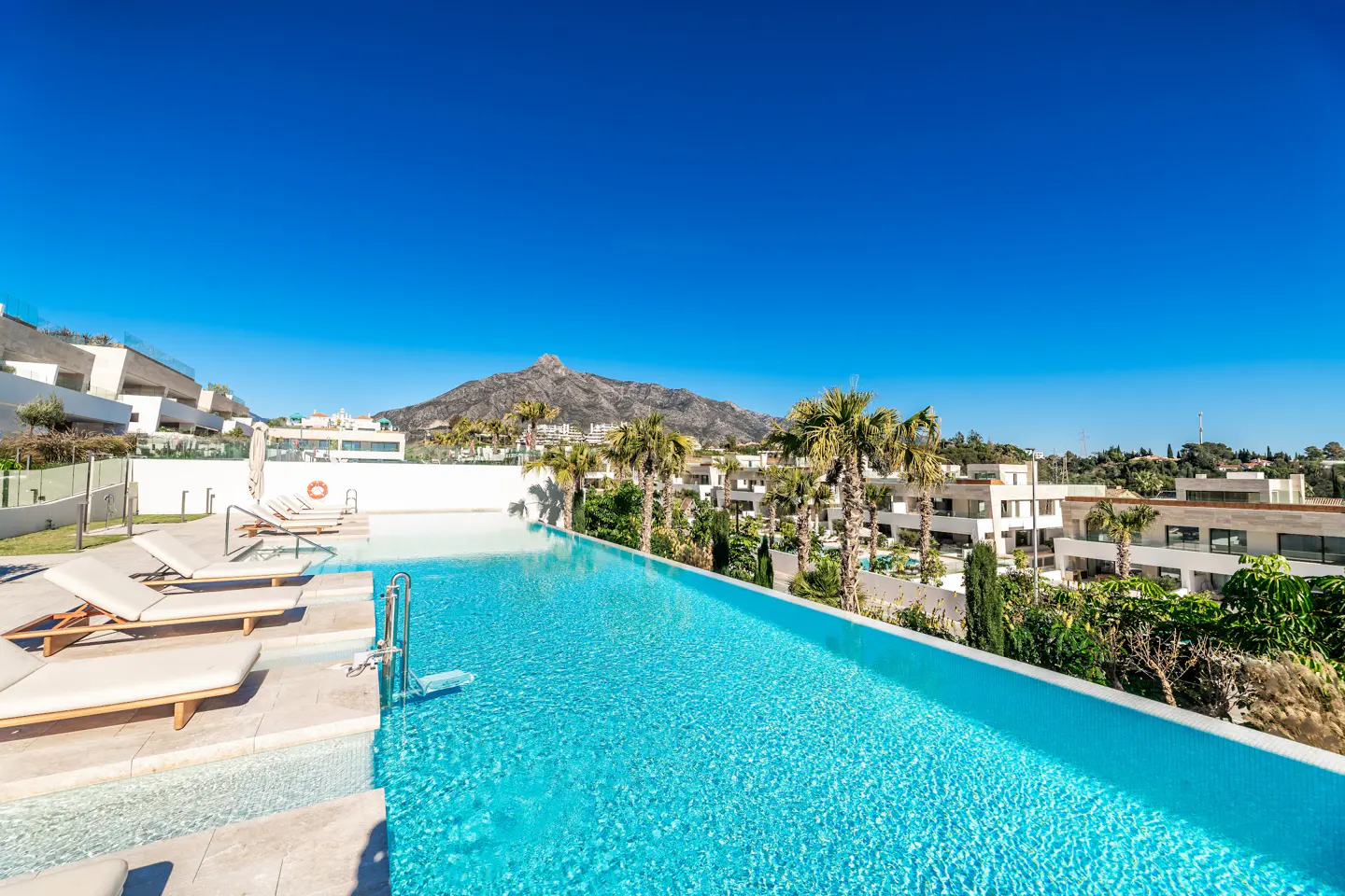 Infinity pool with turquoise water, lounge chairs, and palm trees under a clear blue sky. Mountain in the background.