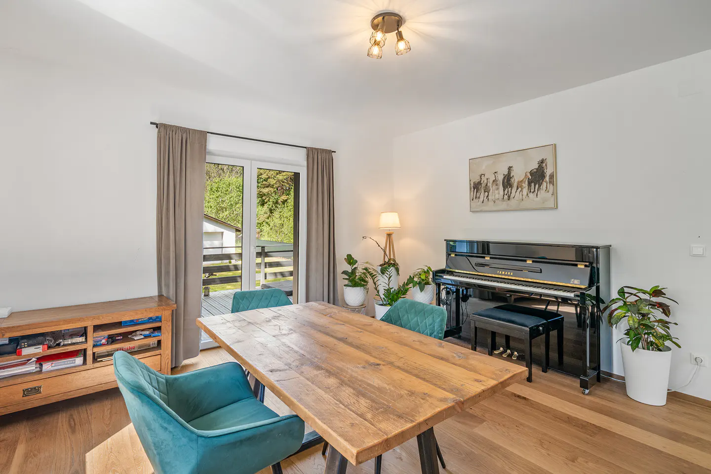 Bright dining room with a wooden table, teal chairs, and a black piano. A door leads to a balcony with green trees in the background.