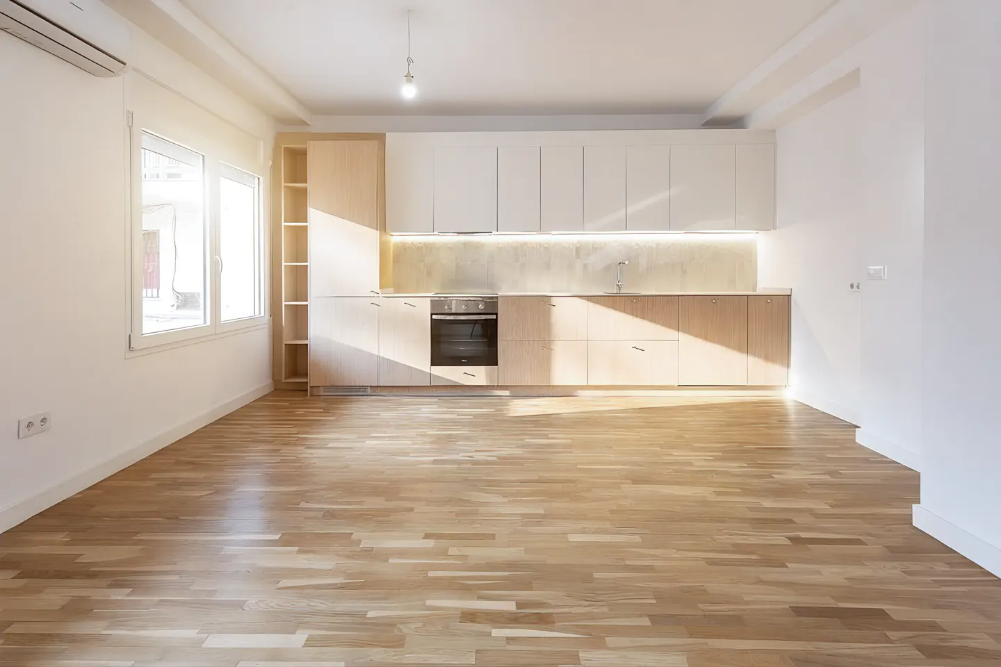 Bright, empty kitchen with light wood floors, white walls, and modern cabinets. Sunlight streams in from a window.