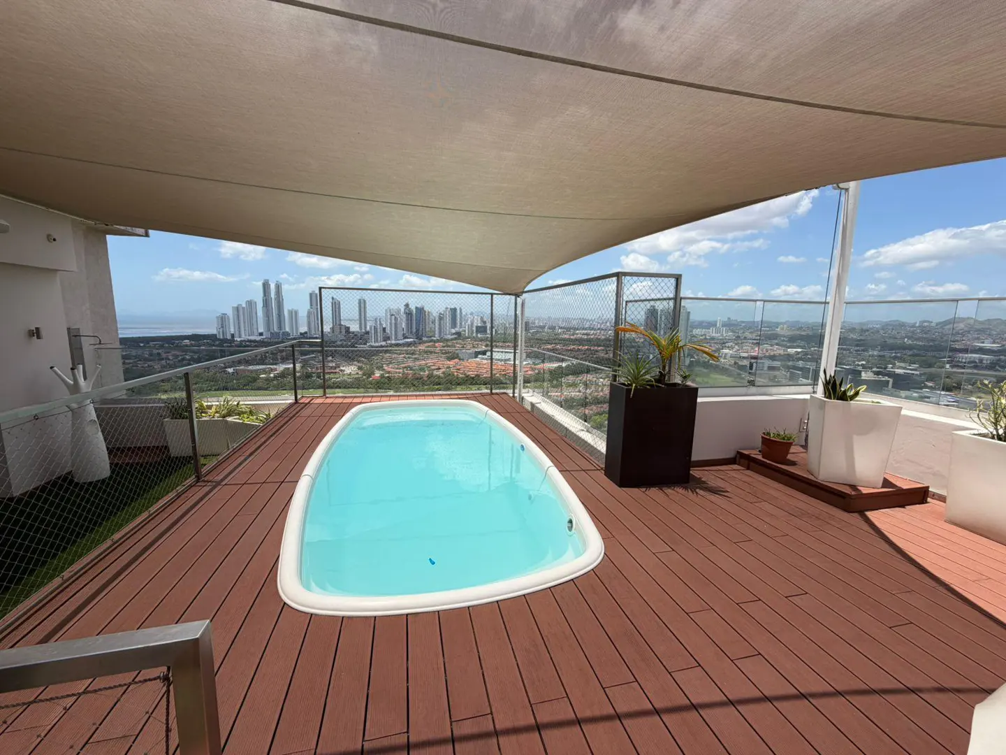 Rooftop deck with a small pool, brown wood flooring, and a beige sunshade. City skyline visible in the background.