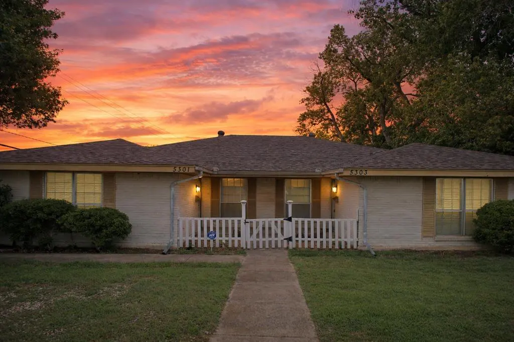 A ranch-style duplex with a white picket fence at sunset. The sky is orange and pink.