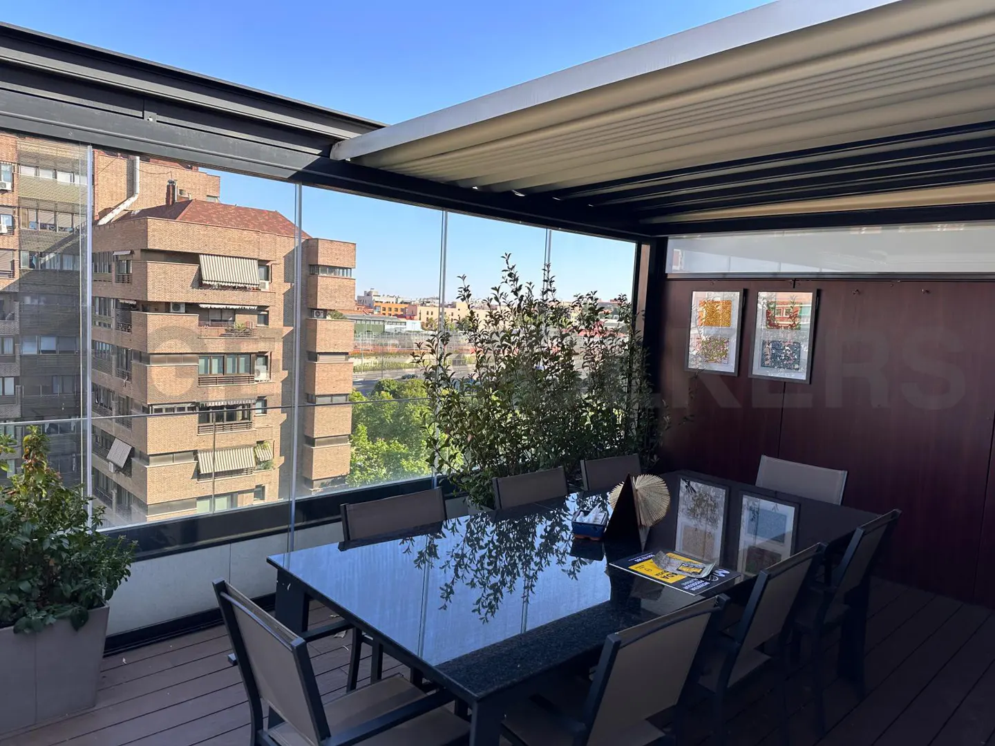 Outdoor dining area with a black table and chairs on a wooden deck, enclosed by glass walls with city views.
