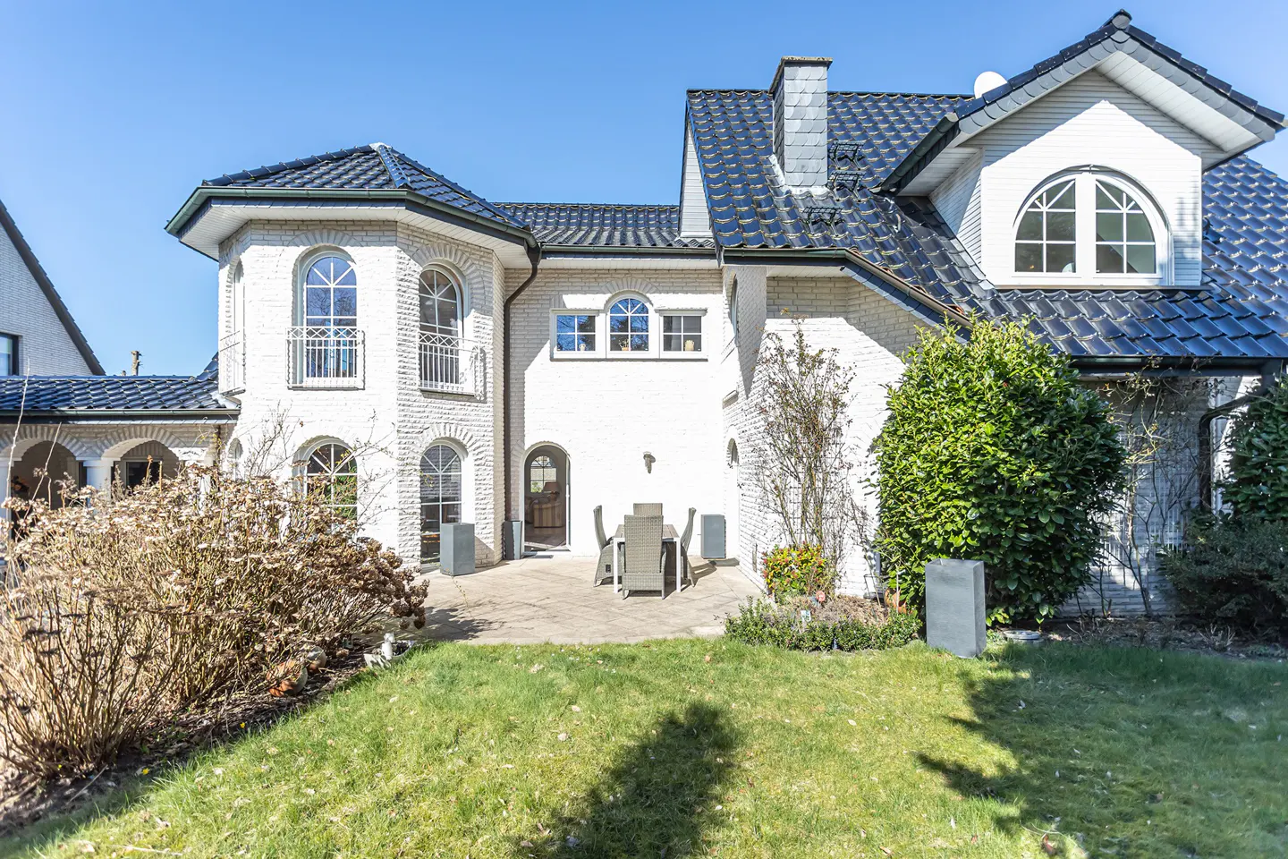 Backyard view of a two-story white brick house with a dark roof and a patio with a table and chairs.