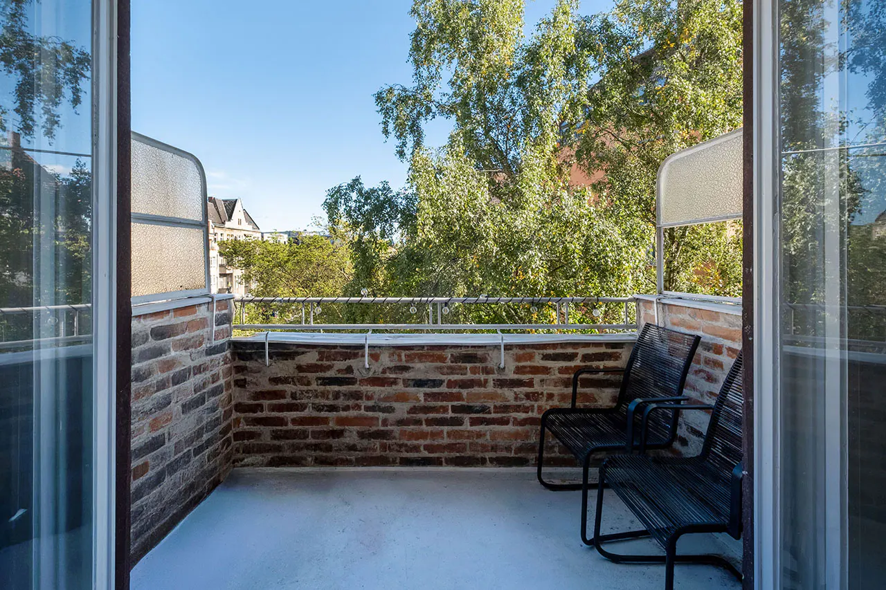 Balcony view with two black chairs, brick wall, and metal railing. Green trees and blue sky visible in the background. Glass doors frame the scene.