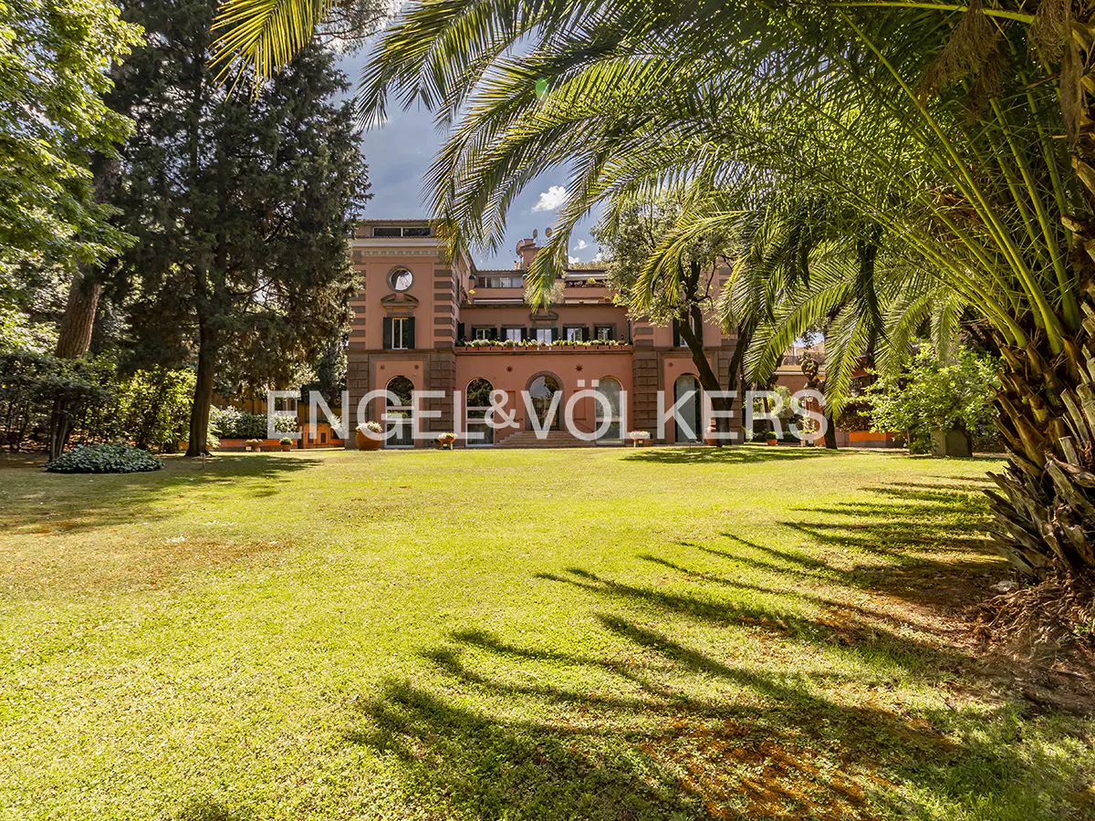 A grand terracotta-colored villa stands behind a lush green lawn, framed by palm trees and tall, leafy garden trees.