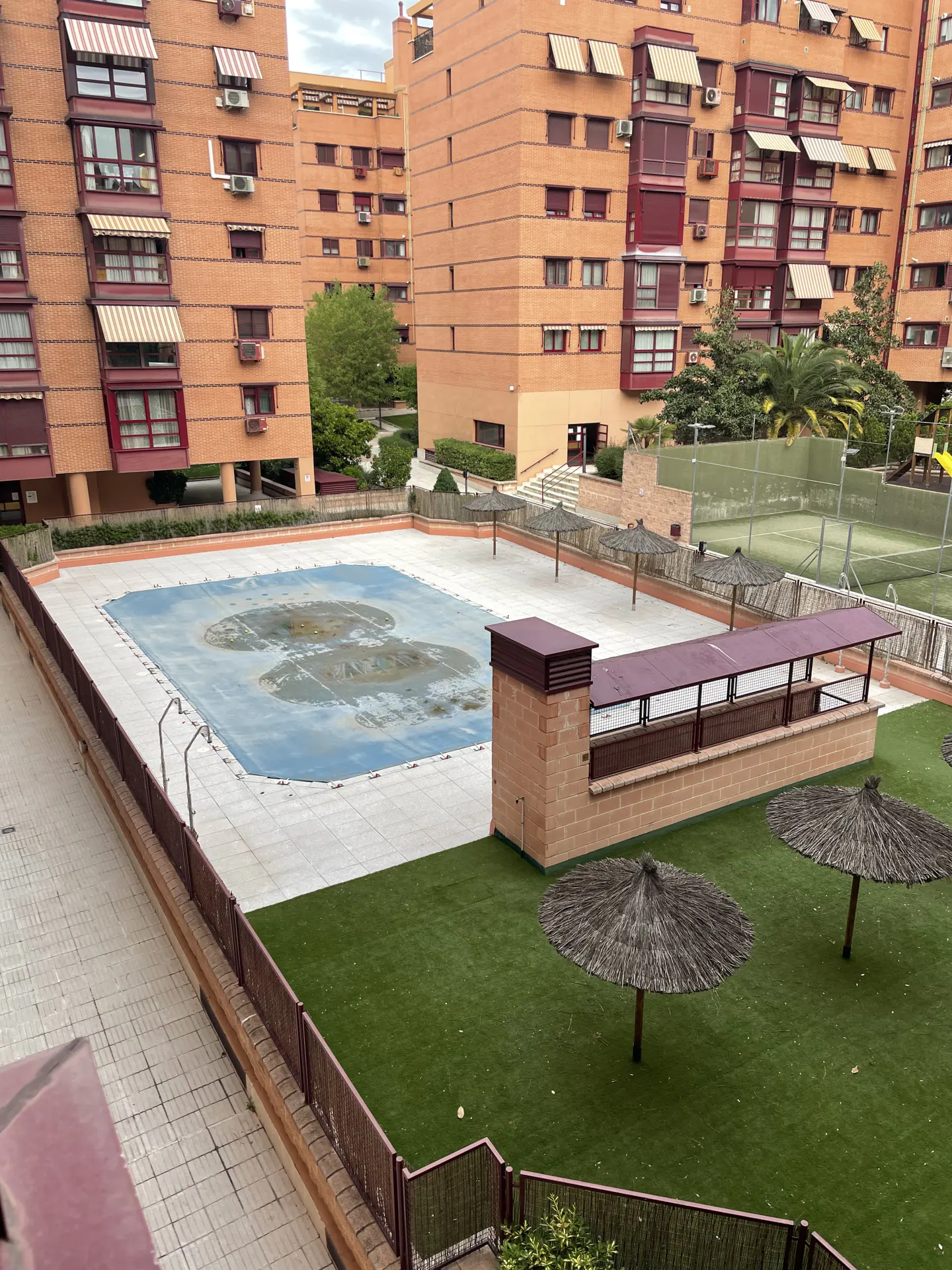 View of a covered pool, green lawn, and brick apartment buildings. There are straw umbrellas and a tennis court in the background.