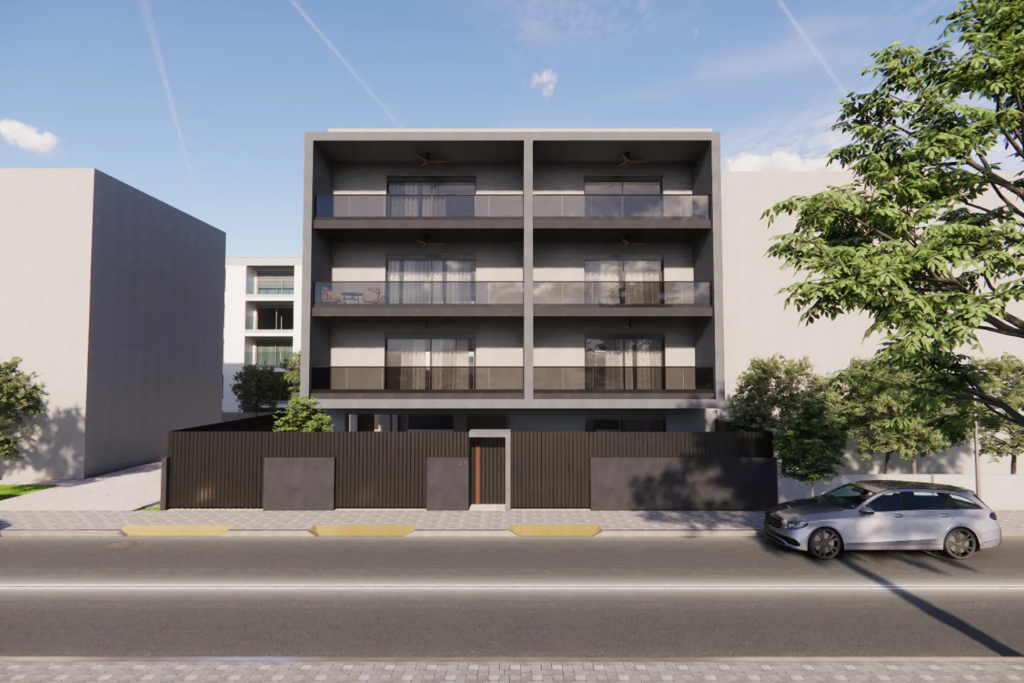 Modern gray apartment building with balconies, behind a dark fence. A silver car is parked on the street.