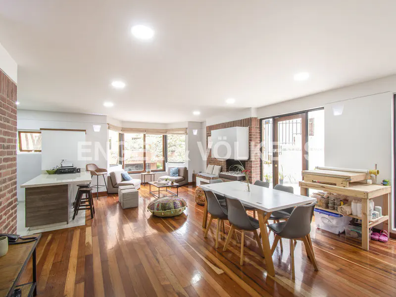Open-concept living space with hardwood floors, brick fireplace, dining table with gray chairs, and a kitchen island. Natural light fills the room.