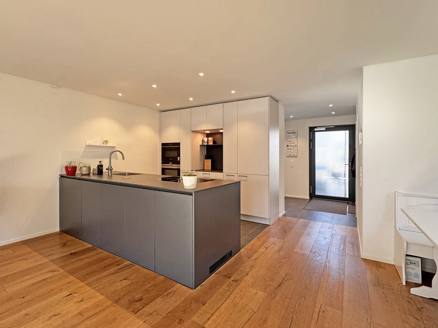 A modern kitchen with wood floors, a gray island, and white cabinets. A black door is visible in the background.