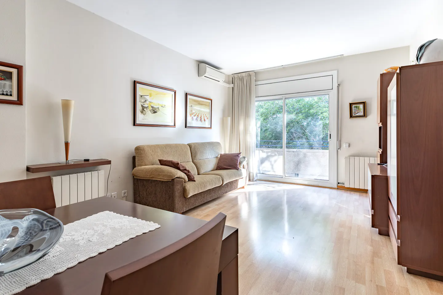 Bright living room with a brown sofa, wood floors, and a dining table. Sliding glass doors lead to a balcony with green trees.