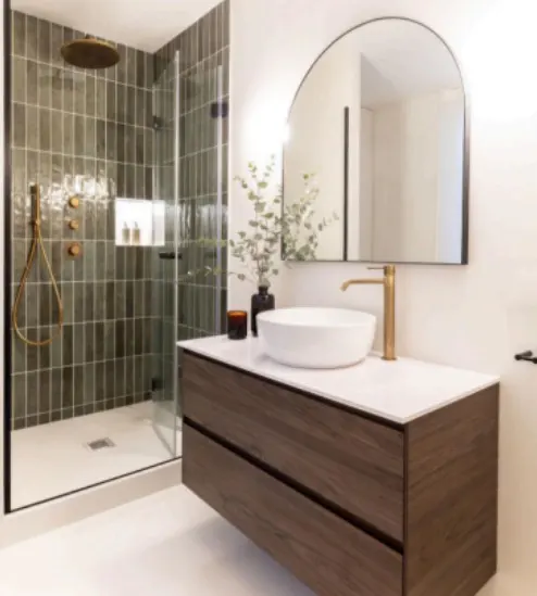 Bathroom with a glass-enclosed shower with gray tiles and gold fixtures, a wood vanity with a white countertop, and an arched mirror.