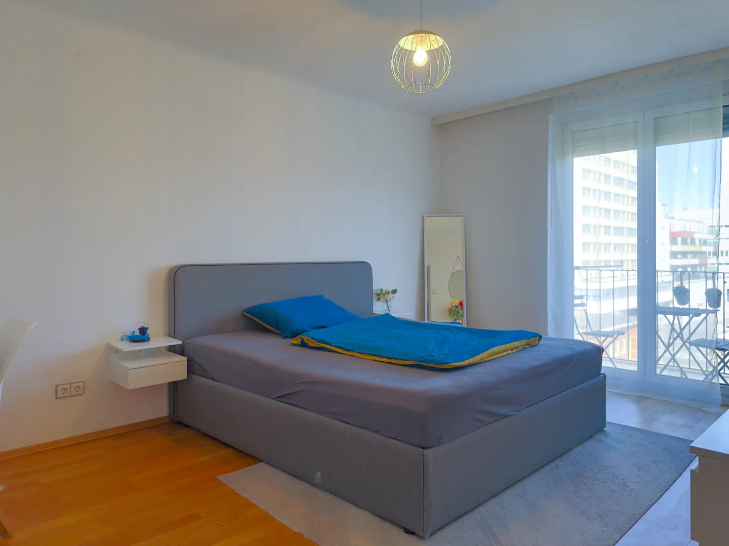 A bright bedroom with a gray bed, blue bedding, and a balcony with a city view. A modern light fixture hangs from the ceiling.