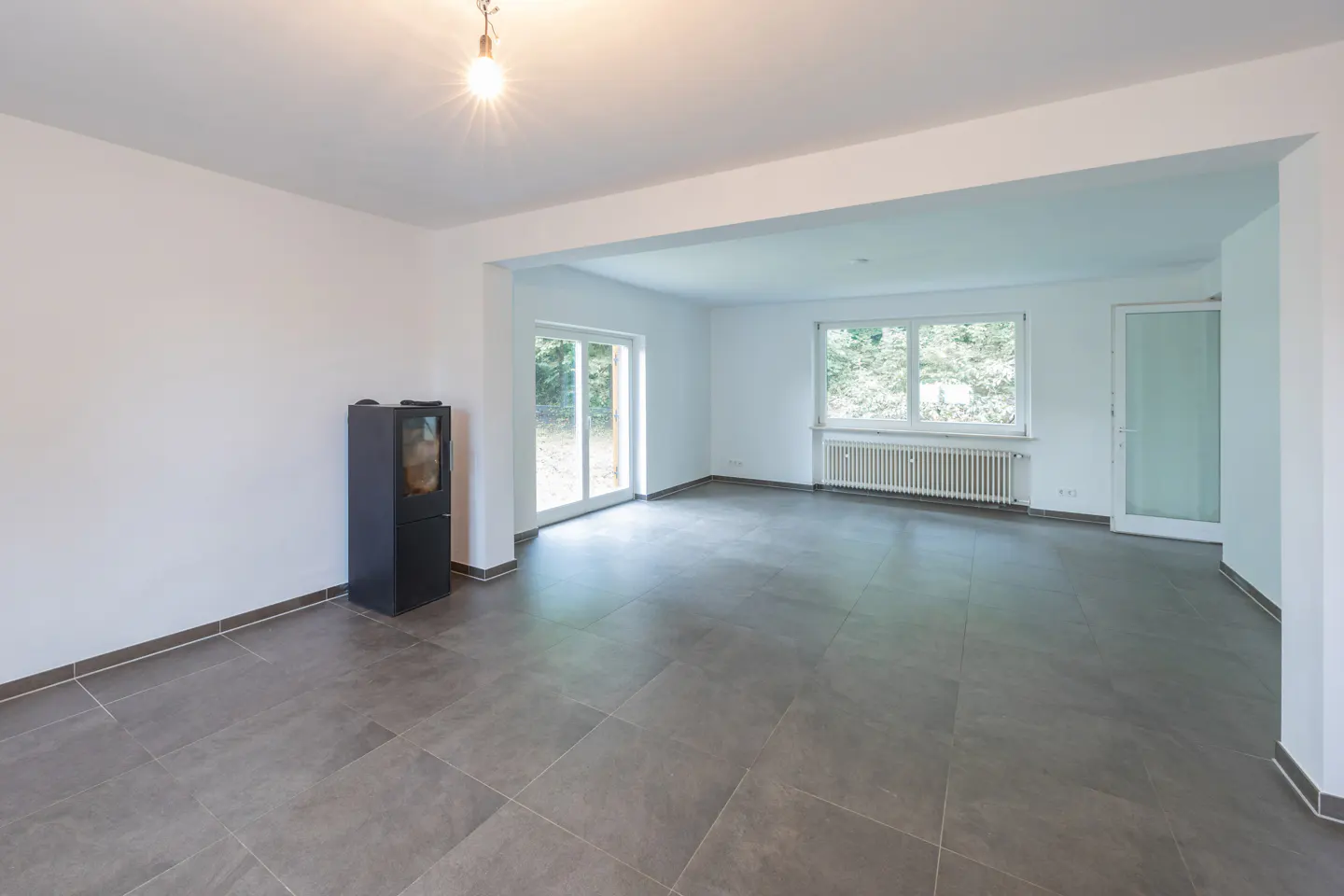 Bright, empty room with gray tile floor, white walls, and a black wood-burning stove. Windows and glass doors offer views of greenery.