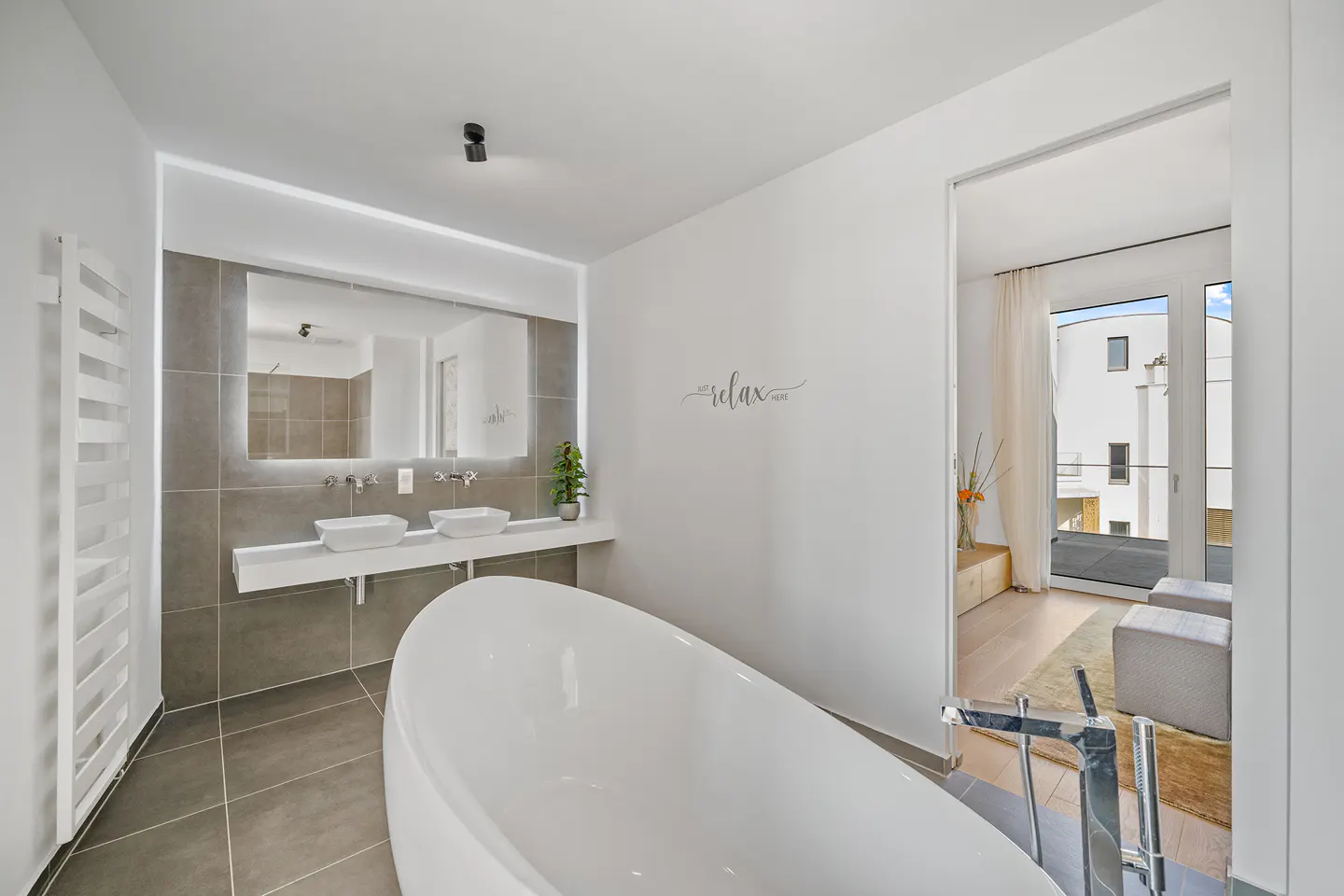 Bright bathroom with gray tile, white walls, and a white tub. A double sink and a door to a balcony are visible.