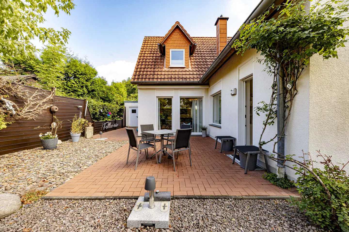 Backyard patio with brick pavers, a table with chairs, and a white house with a red tile roof.