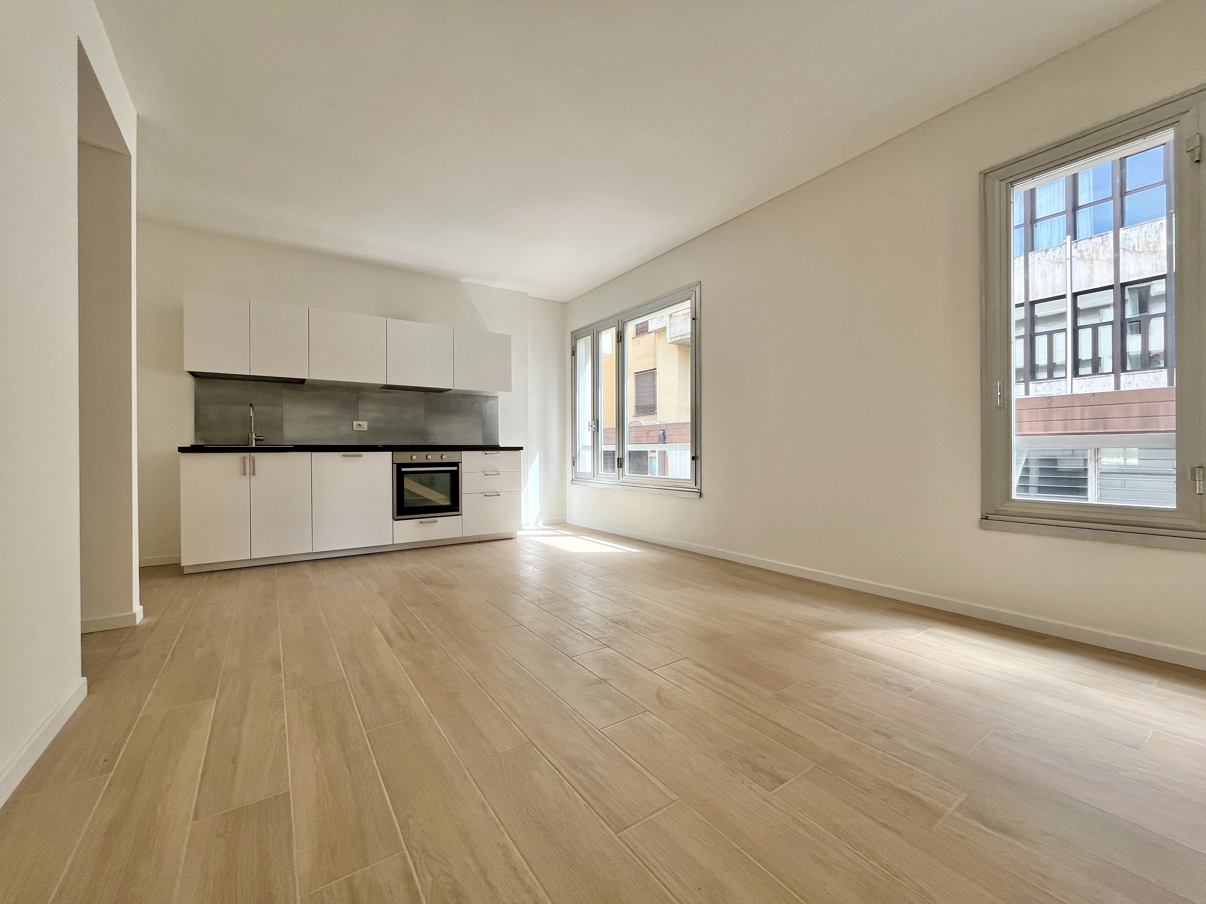 Bright, empty apartment with light wood floors, white walls, and a modern white kitchen. Two windows offer views of buildings outside.