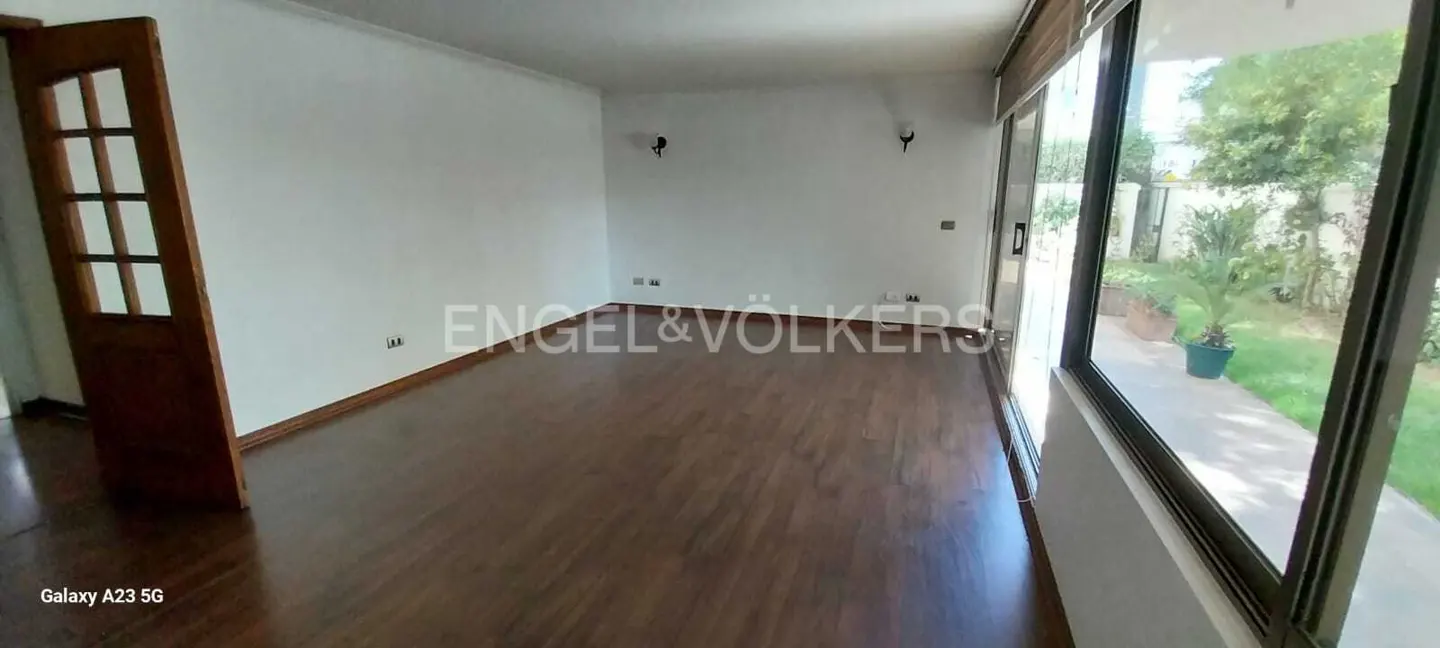 An empty room with dark wood floors, white walls, and a glass door to a garden. The Engel & Volkers logo is in the center.