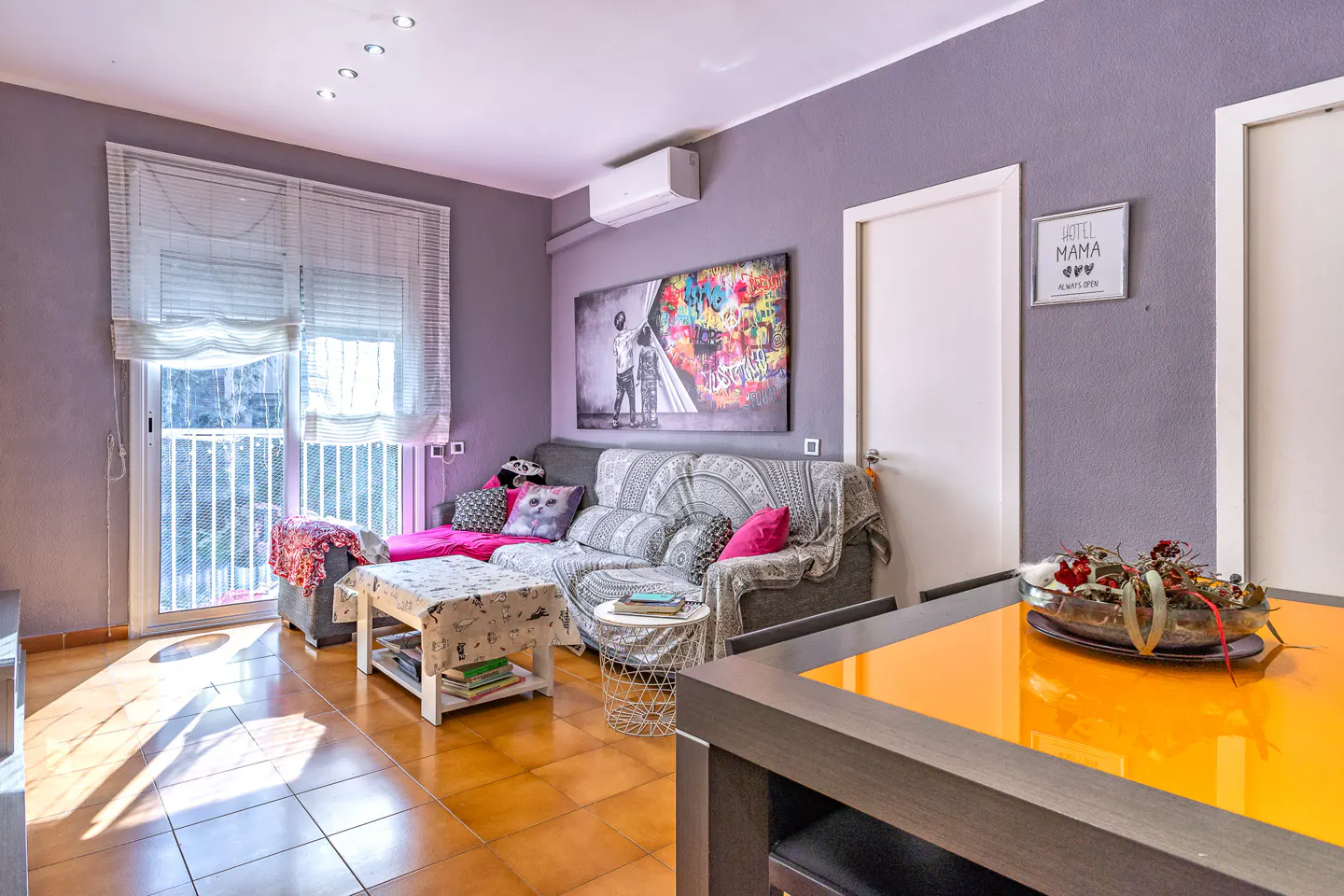 Living room with gray walls, tile floor, and a gray sofa with colorful pillows. A bright orange table is in the foreground.