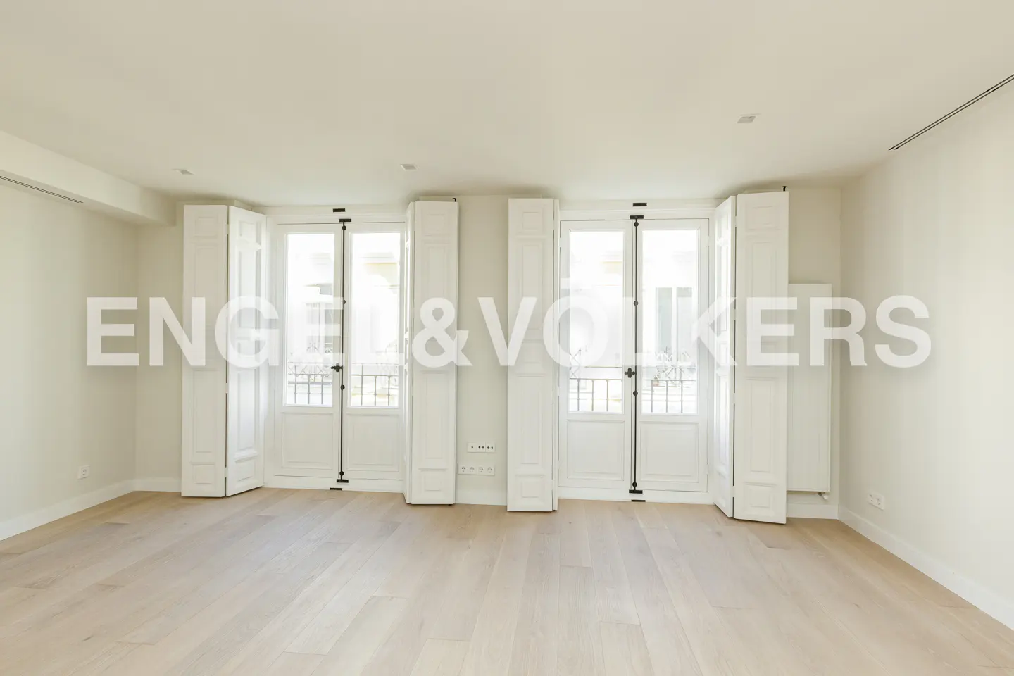 Bright, empty room with light wood floors, white walls, and two sets of tall French doors with white wooden shutters.