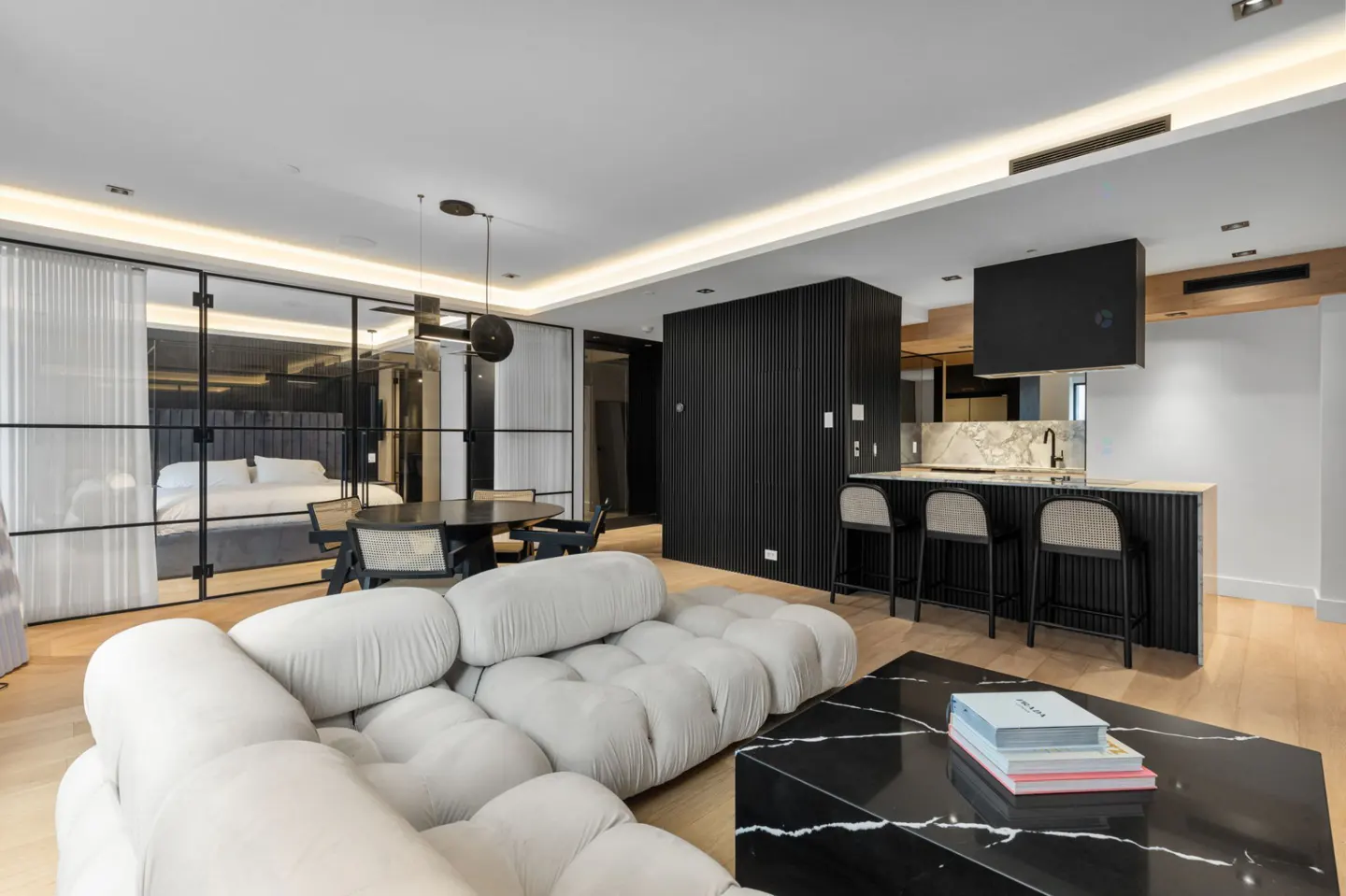 Open-concept living space with a white sofa, black marble coffee table, dining area, and kitchen with bar seating. Bedroom visible through glass doors.