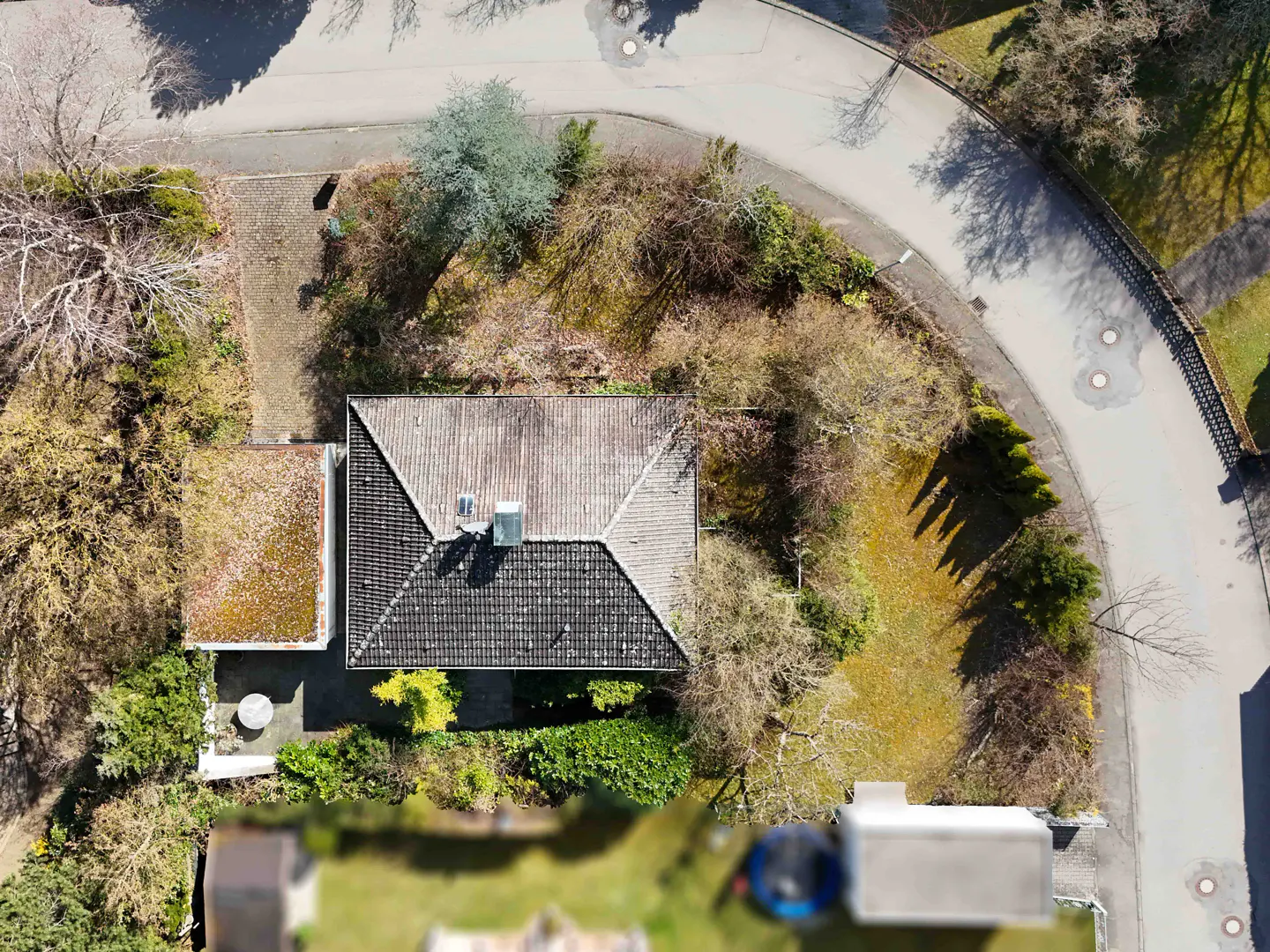 Aerial view of a house with a dark gray tiled roof, surrounded by trees and a curved road.