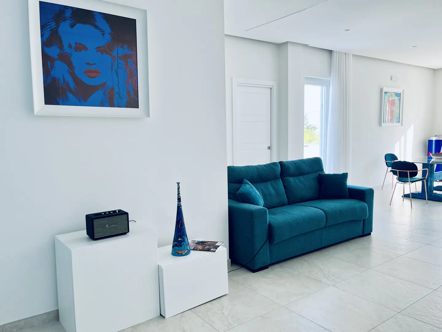 Bright living room with a teal sofa, white walls, and tiled floors. A blue-toned portrait hangs above a white cabinet with a speaker and vase.