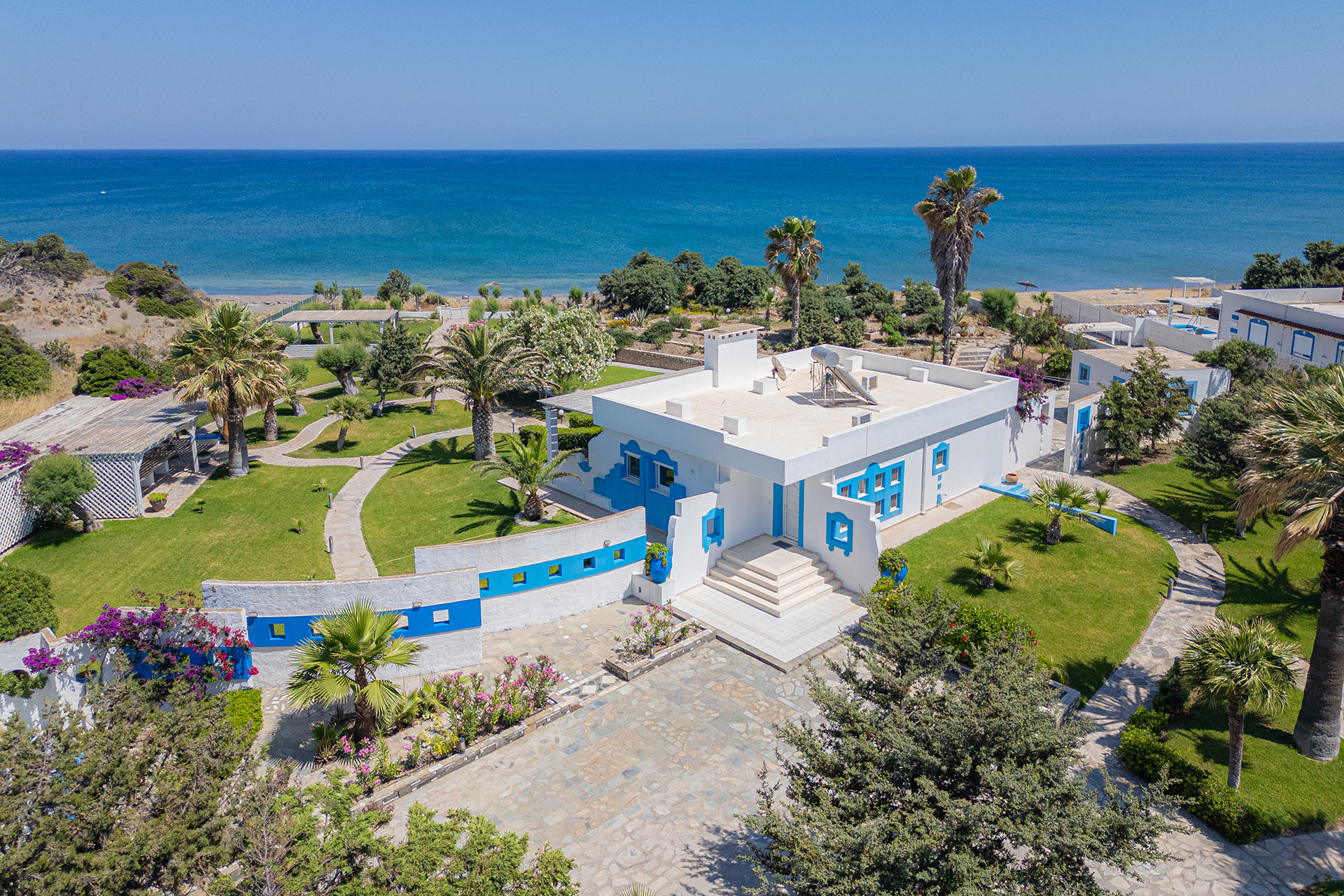 Aerial view of a white villa with blue accents, lush gardens, palm trees, and a stone patio overlooking the blue ocean.