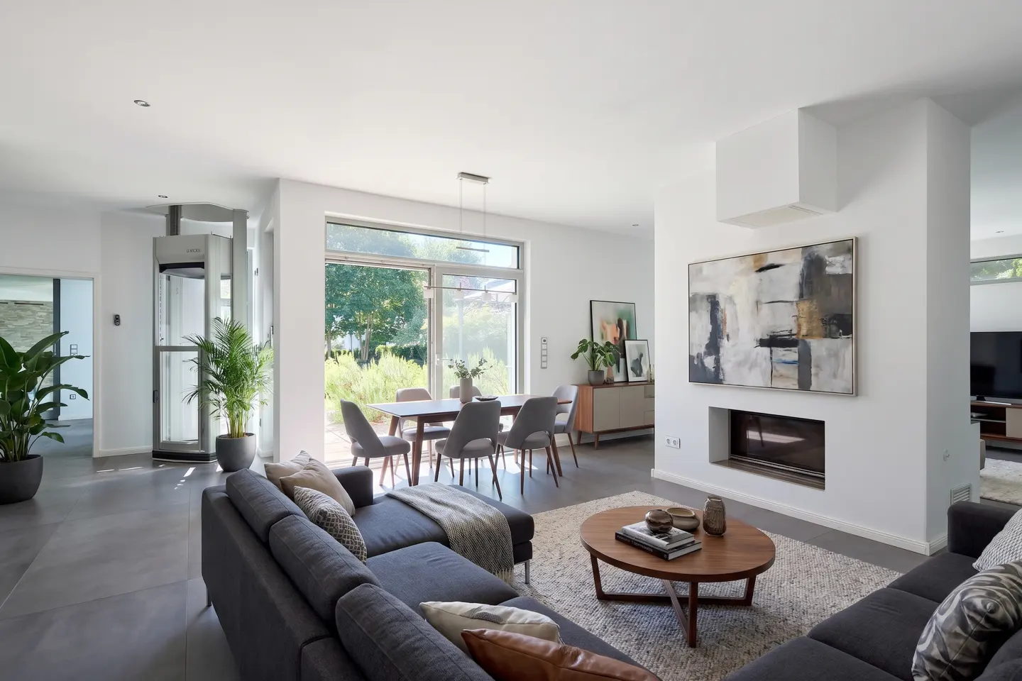 Bright, modern living room with gray sectional sofa, wood table, and dining area. Large windows offer a view of the green outdoors.