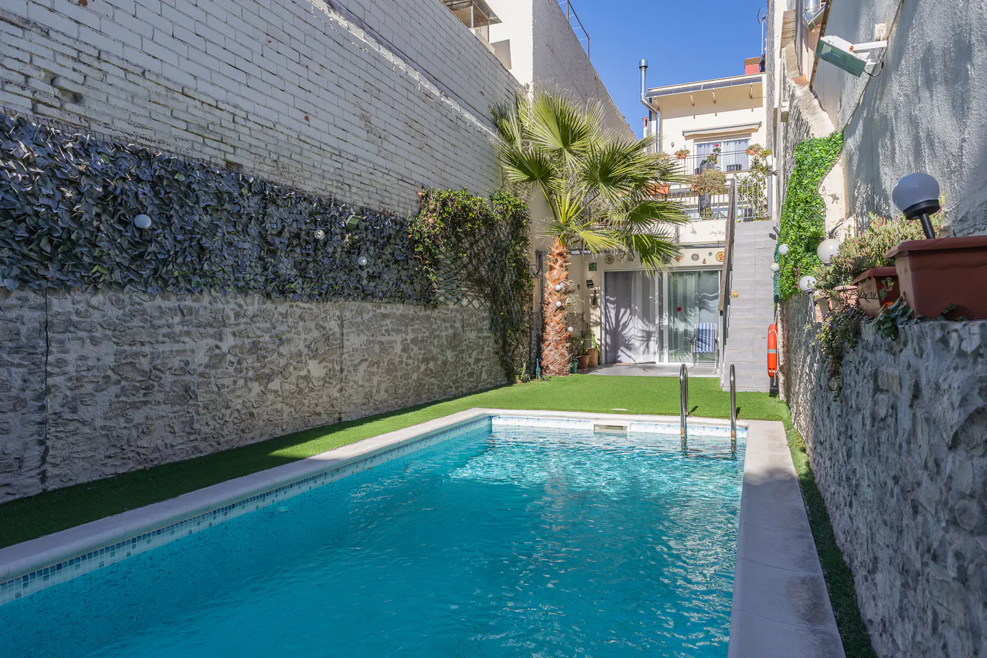 A turquoise pool is surrounded by stone walls, green turf, and a palm tree in a narrow outdoor space.