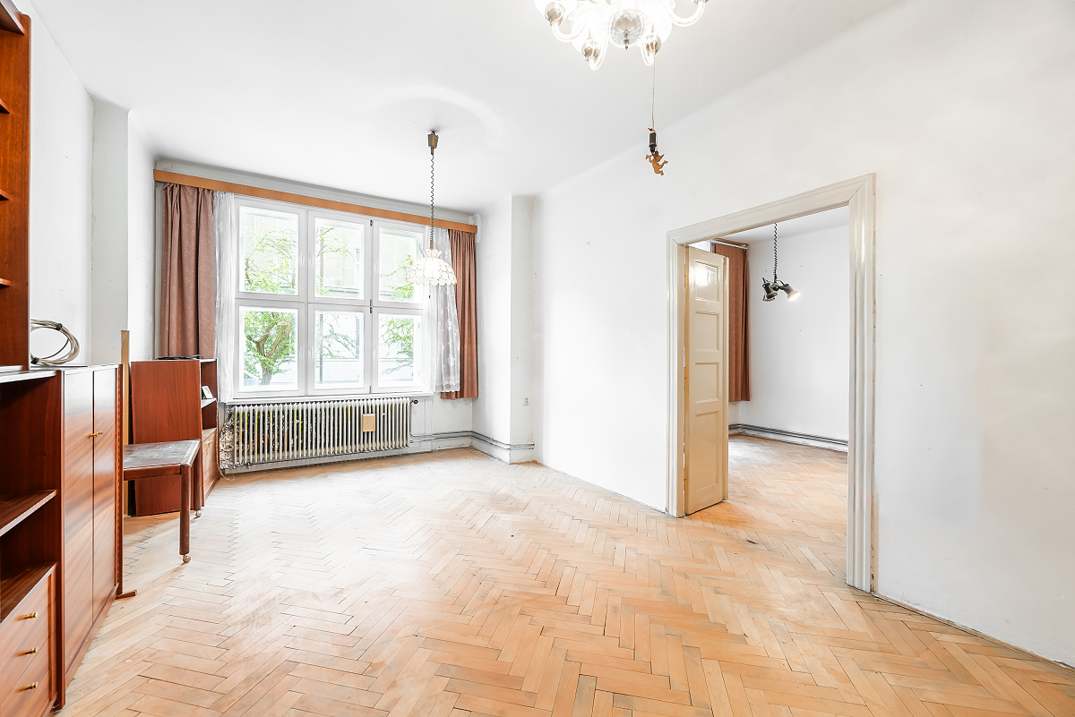 Bright room with herringbone wood floors, white walls, large window with brown curtains, and a doorway to another room. [AI-generated text]