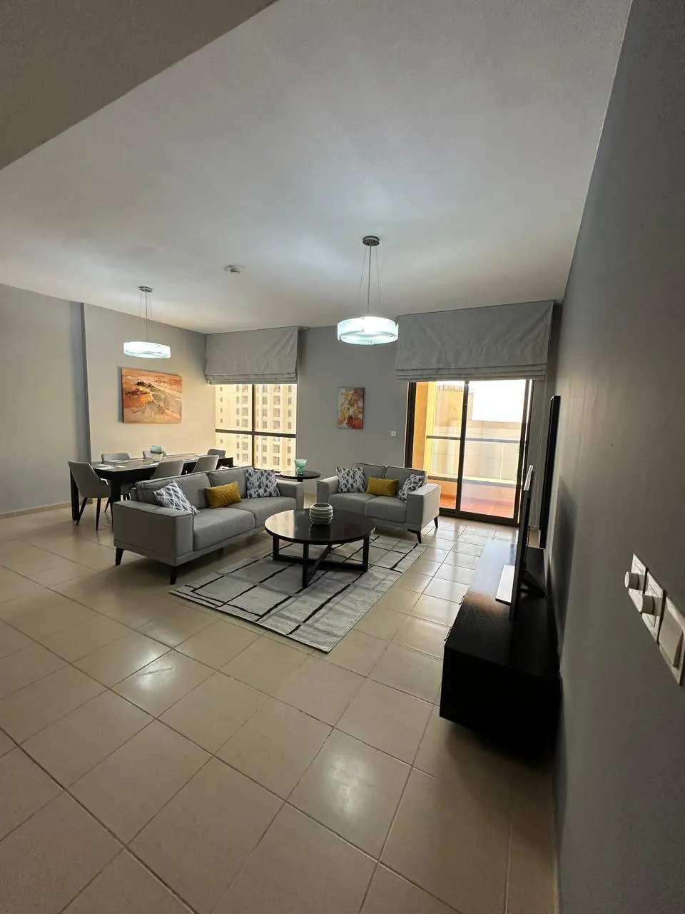 A modern living room with gray sofas, a round coffee table, and a dining area with a table and chairs. Balcony view.