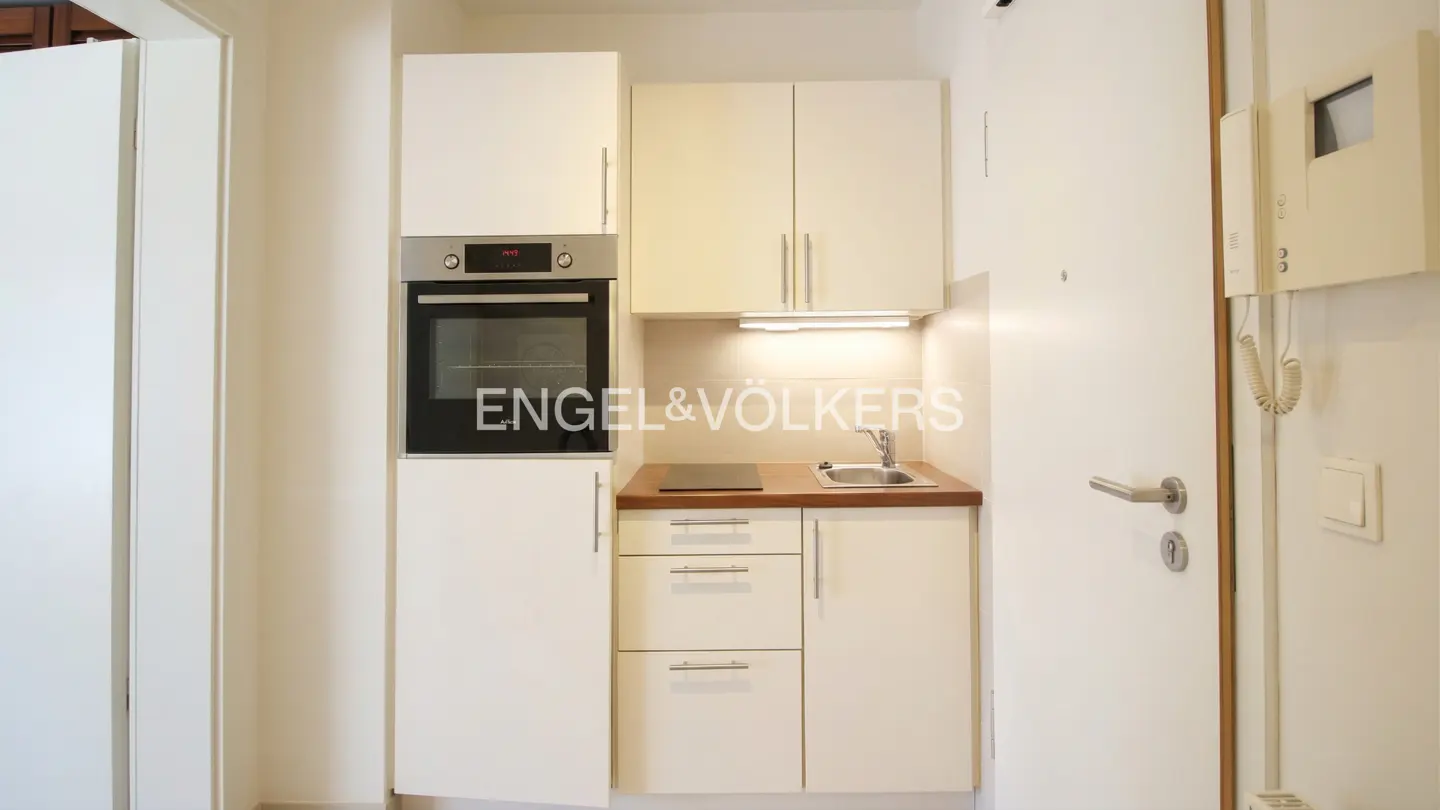 A compact kitchen with white cabinets, a built-in oven, a small sink, and a wooden countertop. A white door with an intercom system is visible on the right.
