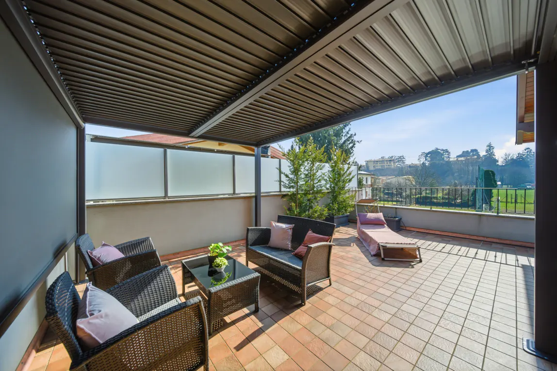 Covered terrace with wicker furniture, a glass coffee table, and a lounge chair overlooking a sunny landscape.