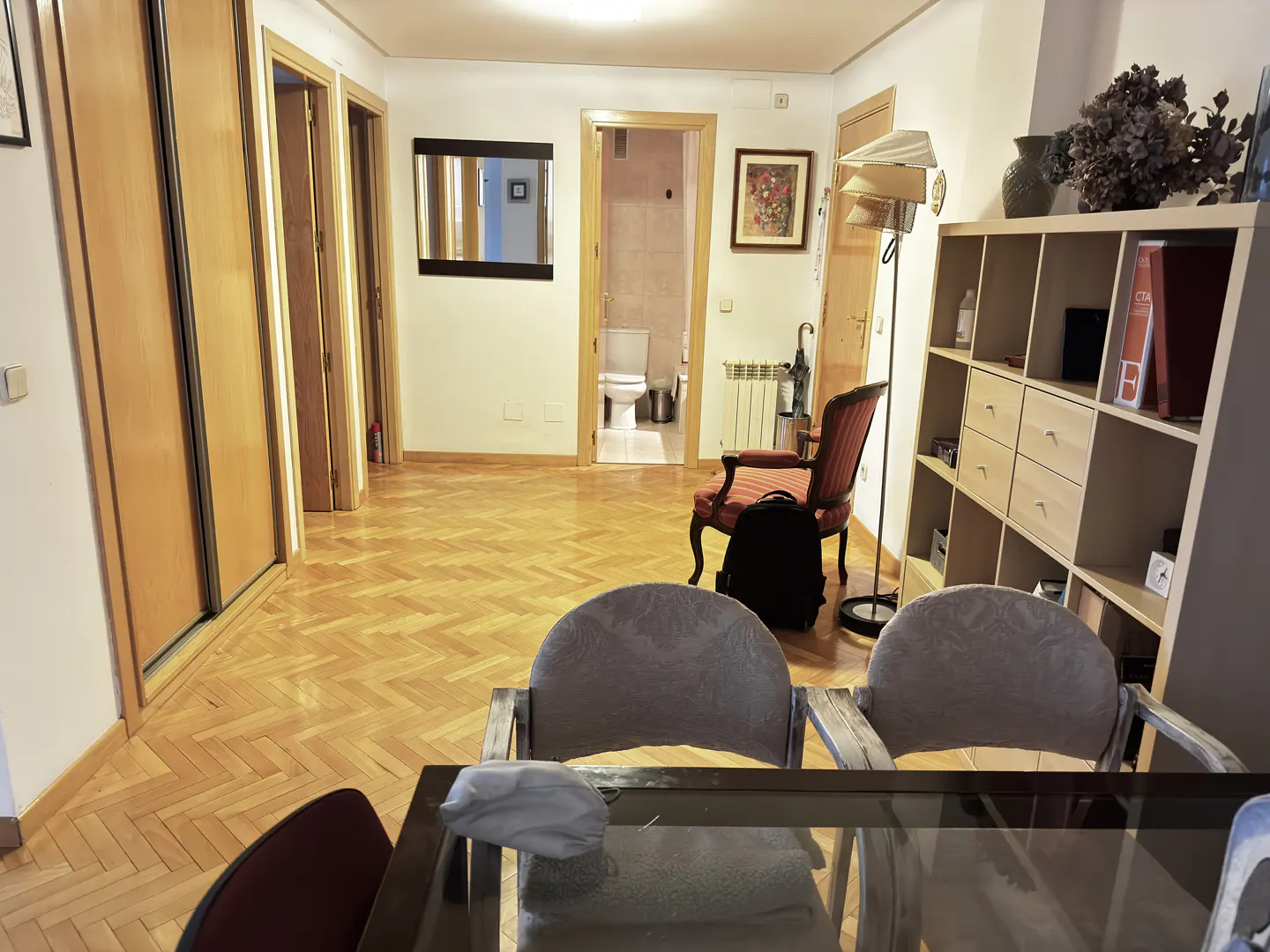 Living room with parquet floor, chairs, bookcase, and a glimpse into a bathroom with a toilet.