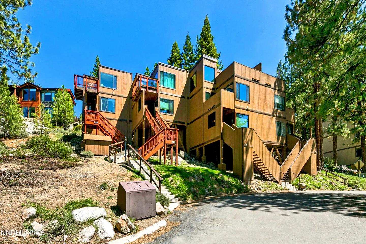 Exterior view of a multi-level, wood-sided condo building with red stairs and balconies, set against a blue sky and green trees.