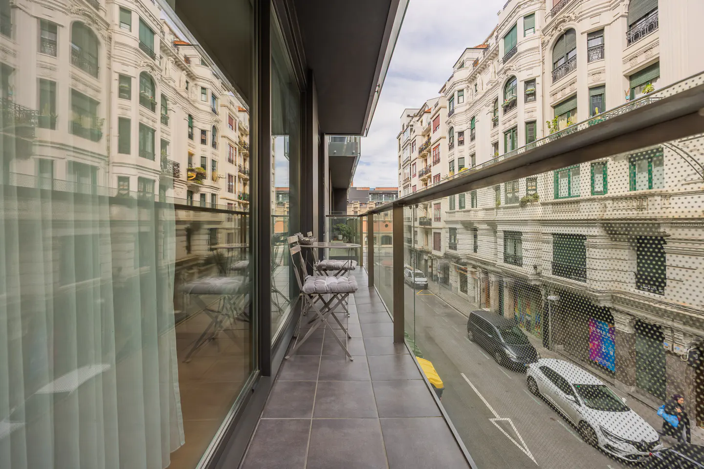 Balcony view of a European city street. Two chairs and a small table sit on the gray tiled balcony with a glass railing. Buildings line the street below.