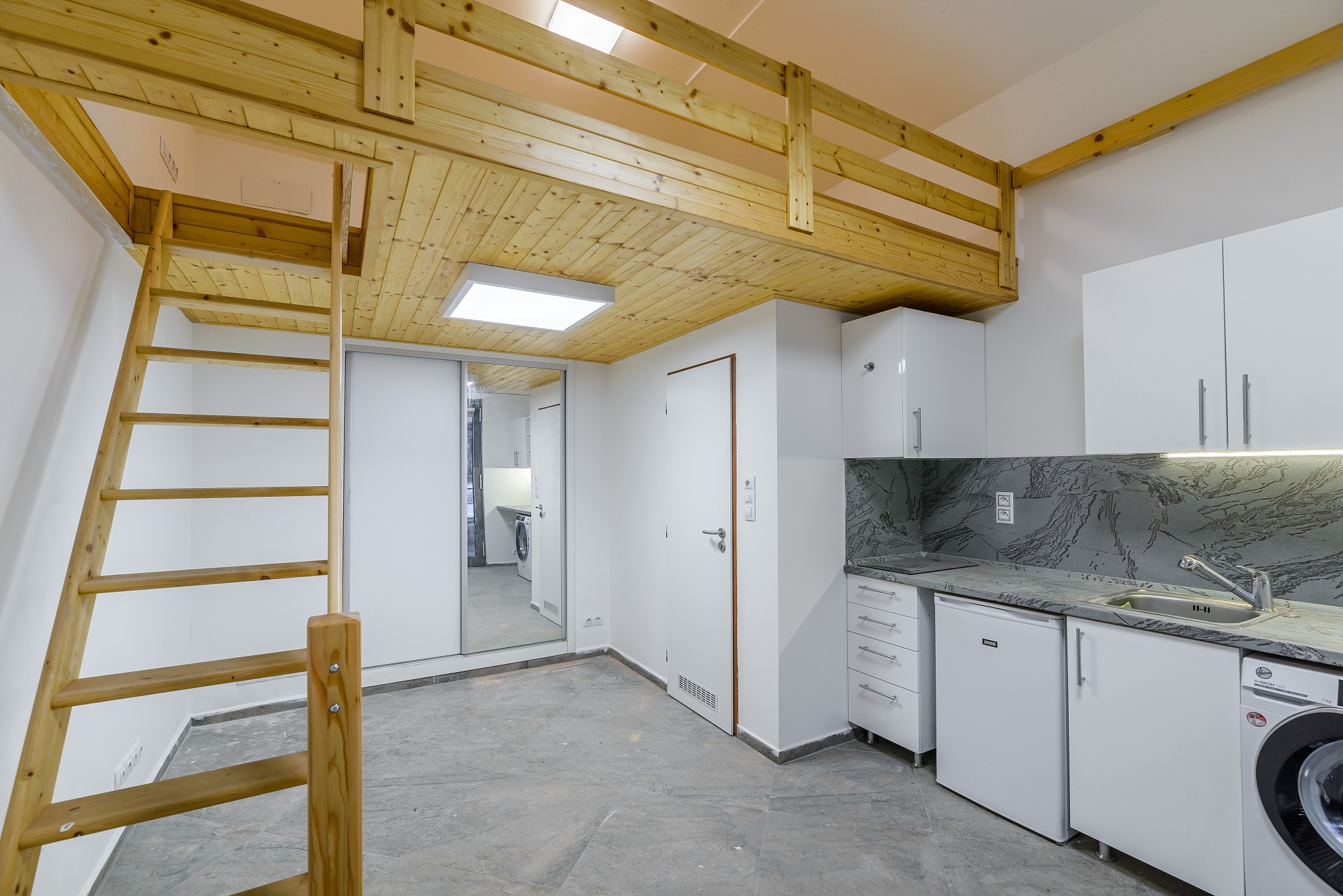 Small studio apartment with a wooden loft bed, ladder, white kitchen cabinets, and a grey stone-patterned backsplash. [AI-generated text]