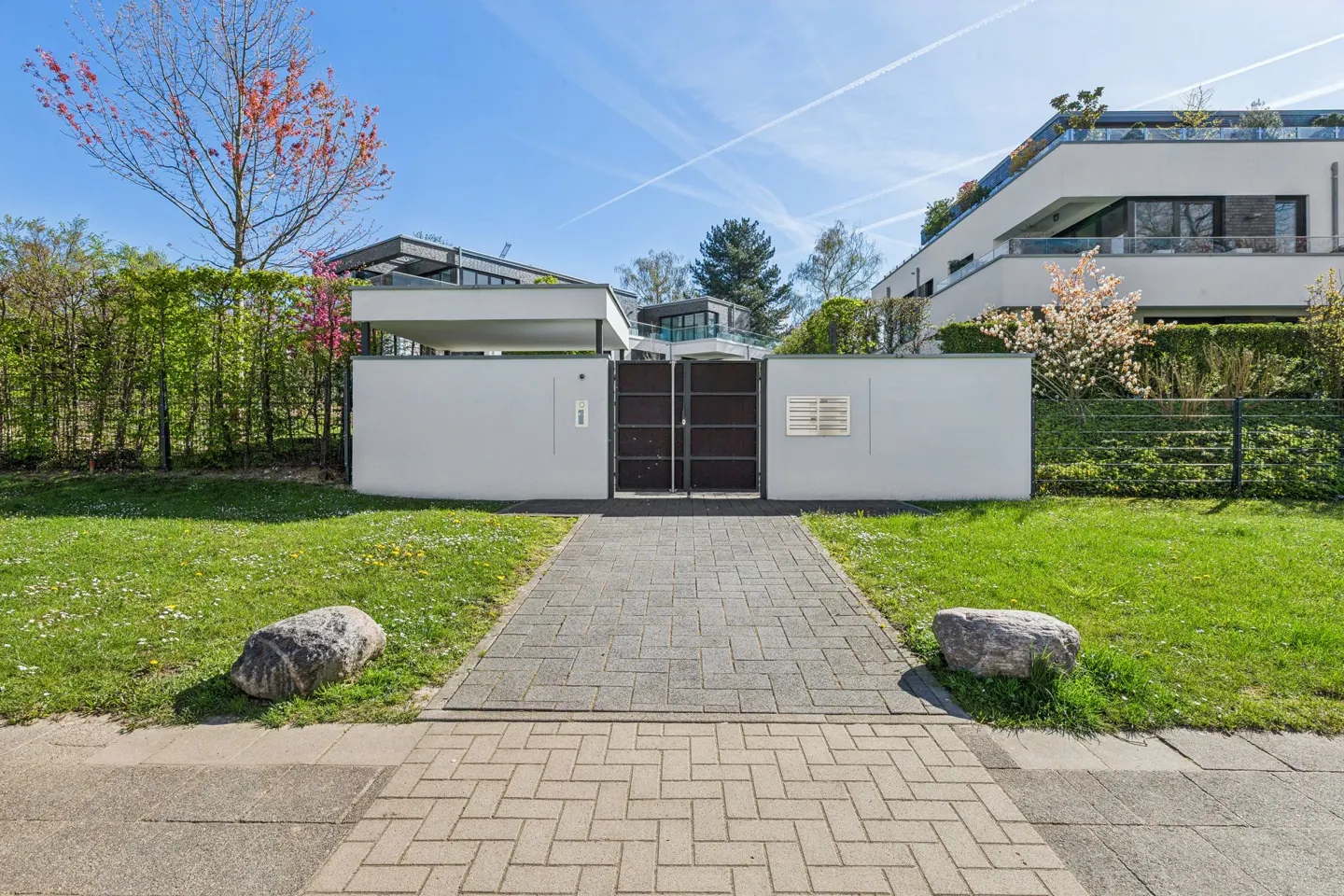 Exterior view of a modern home with a gray brick walkway leading to a black metal gate in a white wall. Green lawn and trees surround the property.