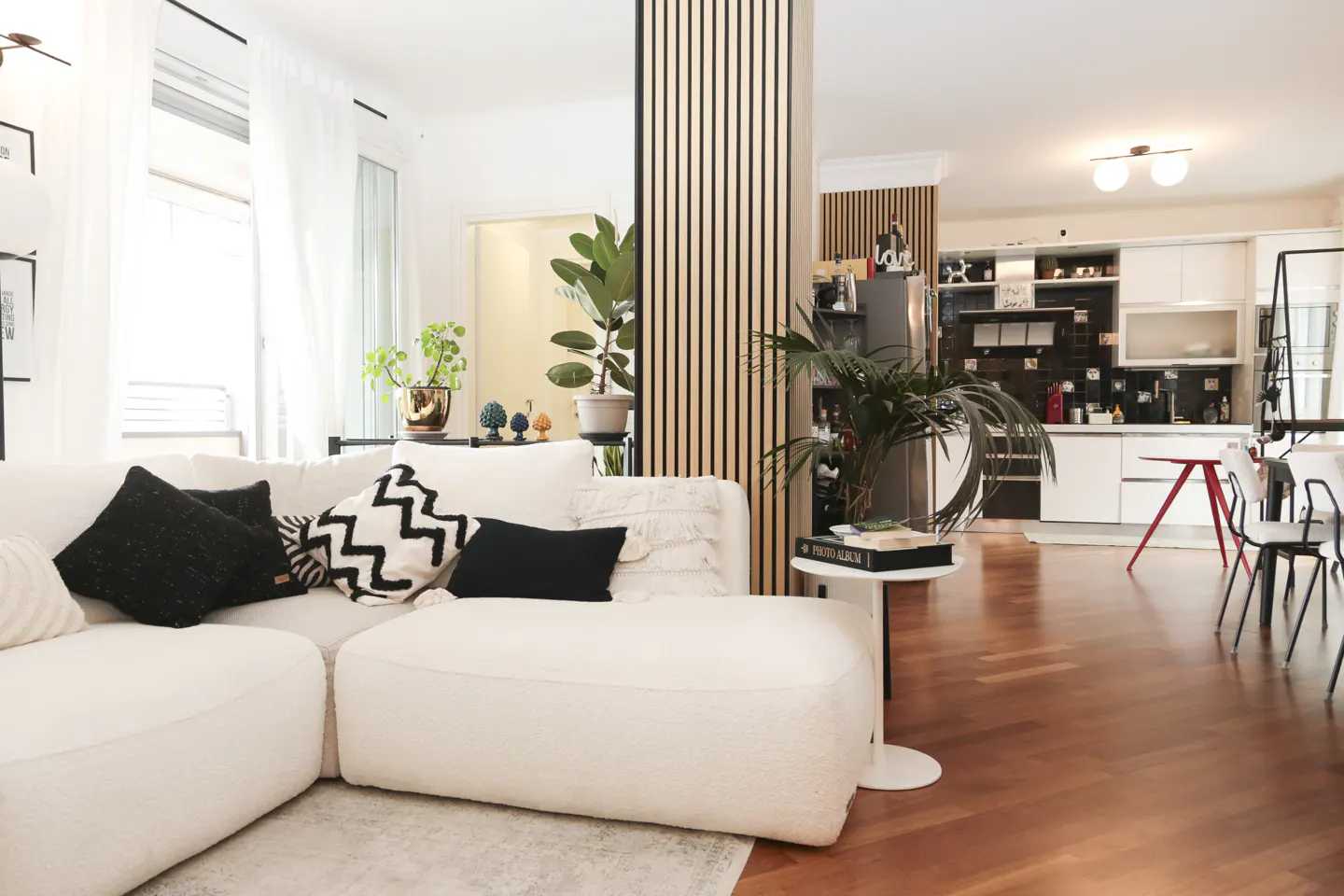 Bright, modern living room with a white couch, black pillows, and wood floors. An open kitchen is visible in the background.