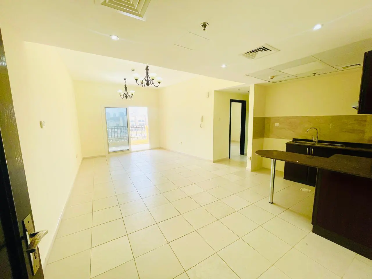 A bright, empty apartment with cream walls, tile floors, a kitchen counter, and a balcony. Chandeliers hang from the ceiling.