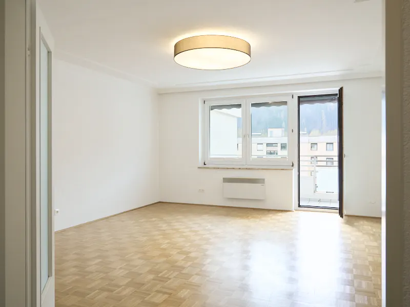 Bright, empty room with parquet floors, white walls, and a large window. A round ceiling light illuminates the space. Balcony access is visible.