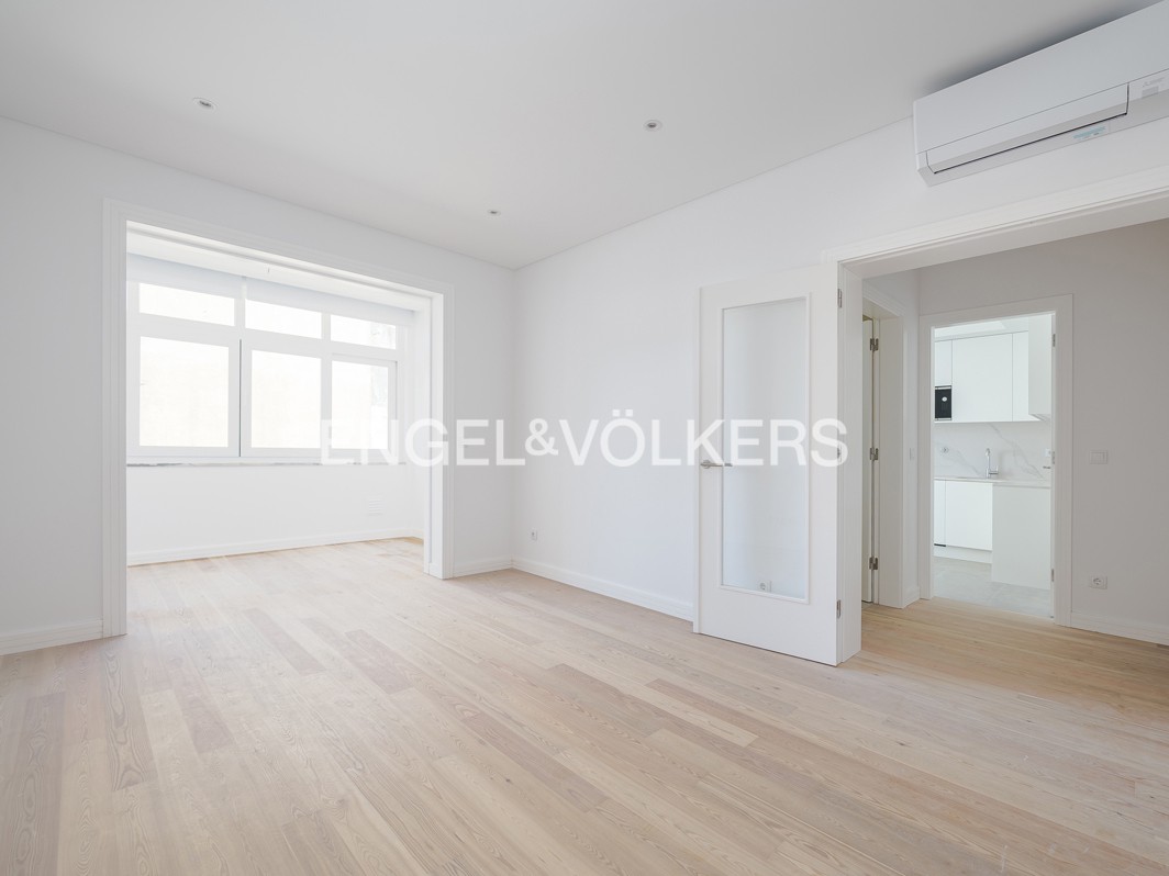 Bright, empty room with light wood floors, white walls, and a window. A doorway leads to a white kitchen. Air conditioning unit on the wall.