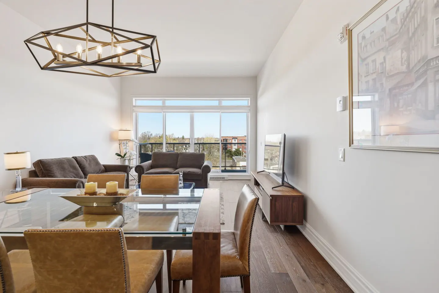 Bright living room with brown sofas, a glass table with leather chairs, and a modern chandelier. A large window offers a view of the outside.