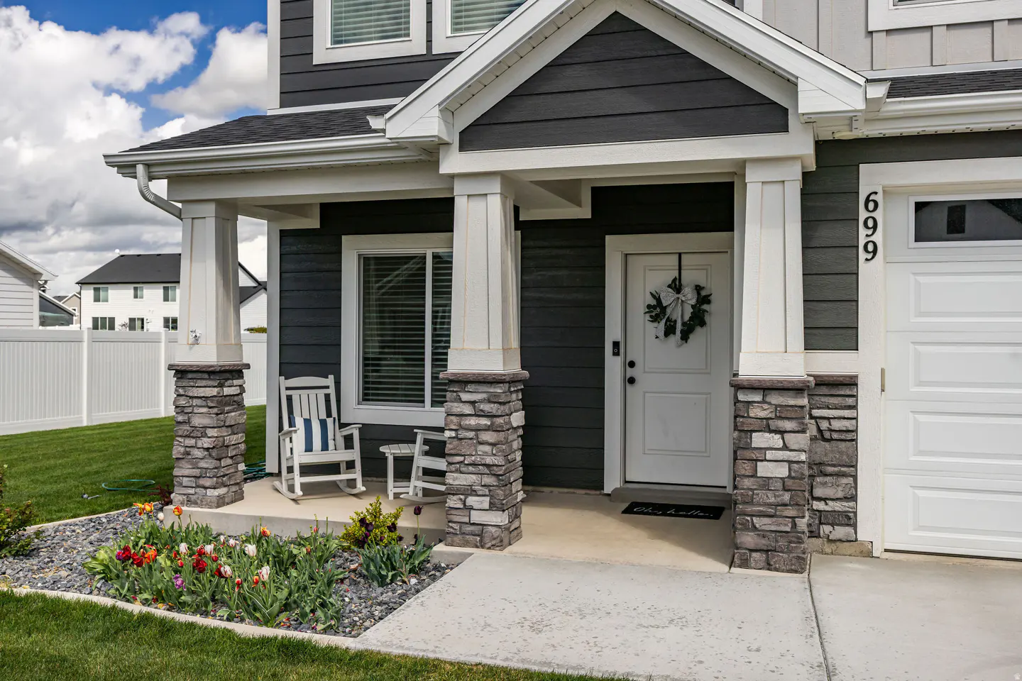 Front view of a two-story house with gray siding, white trim, and a white front door with a wreath. A white rocking chair sits on the porch.