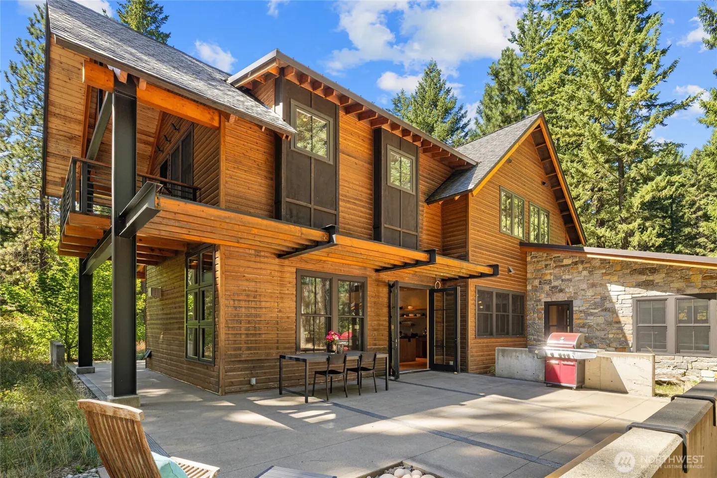 Modern wood-sided home with a stone accent wall, concrete patio, outdoor dining set, and red grill in a forest setting.