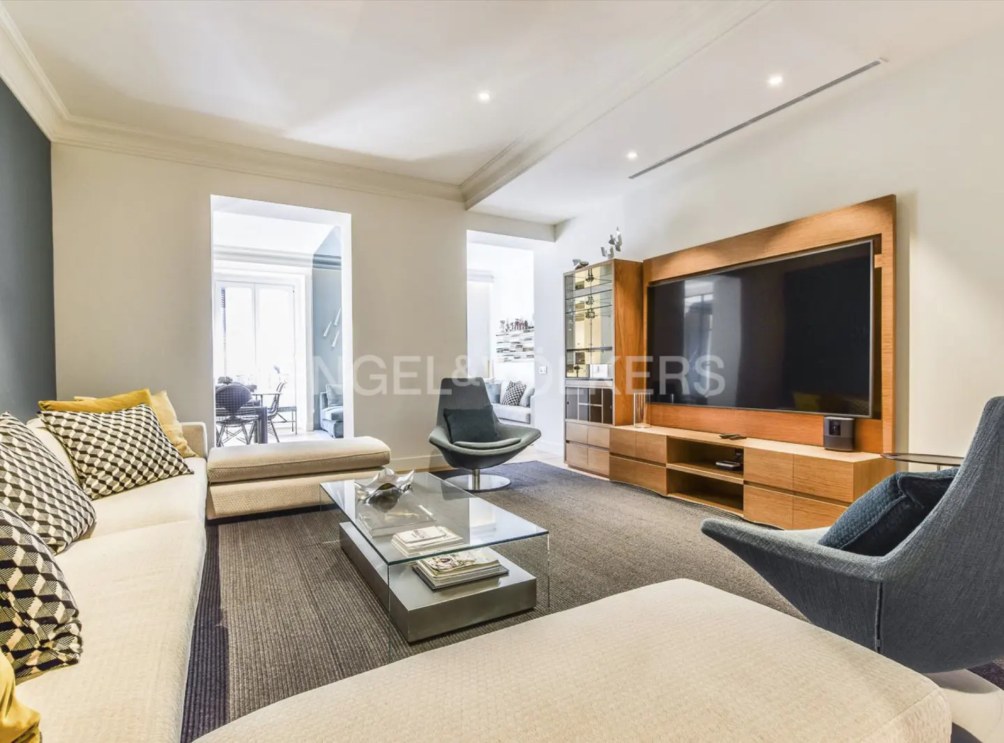 Living room with white sofas, glass table, and a large TV mounted on a wooden entertainment center. Gray carpet and blue accent chairs.