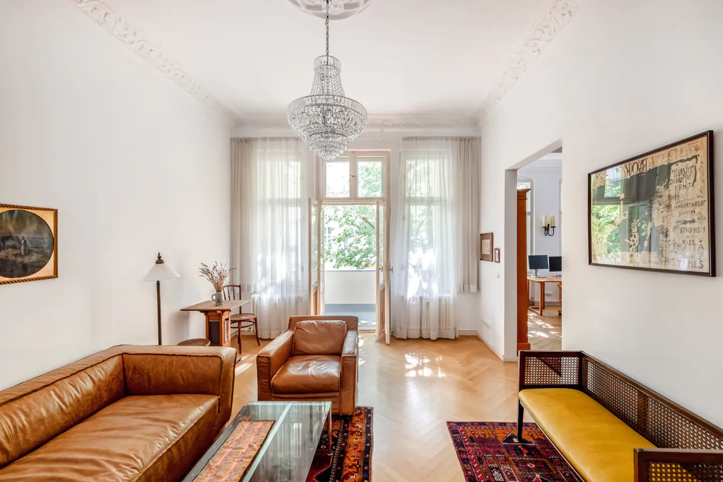 Bright living room with herringbone wood floors, leather sofa, armchair, glass coffee table, and a crystal chandelier.